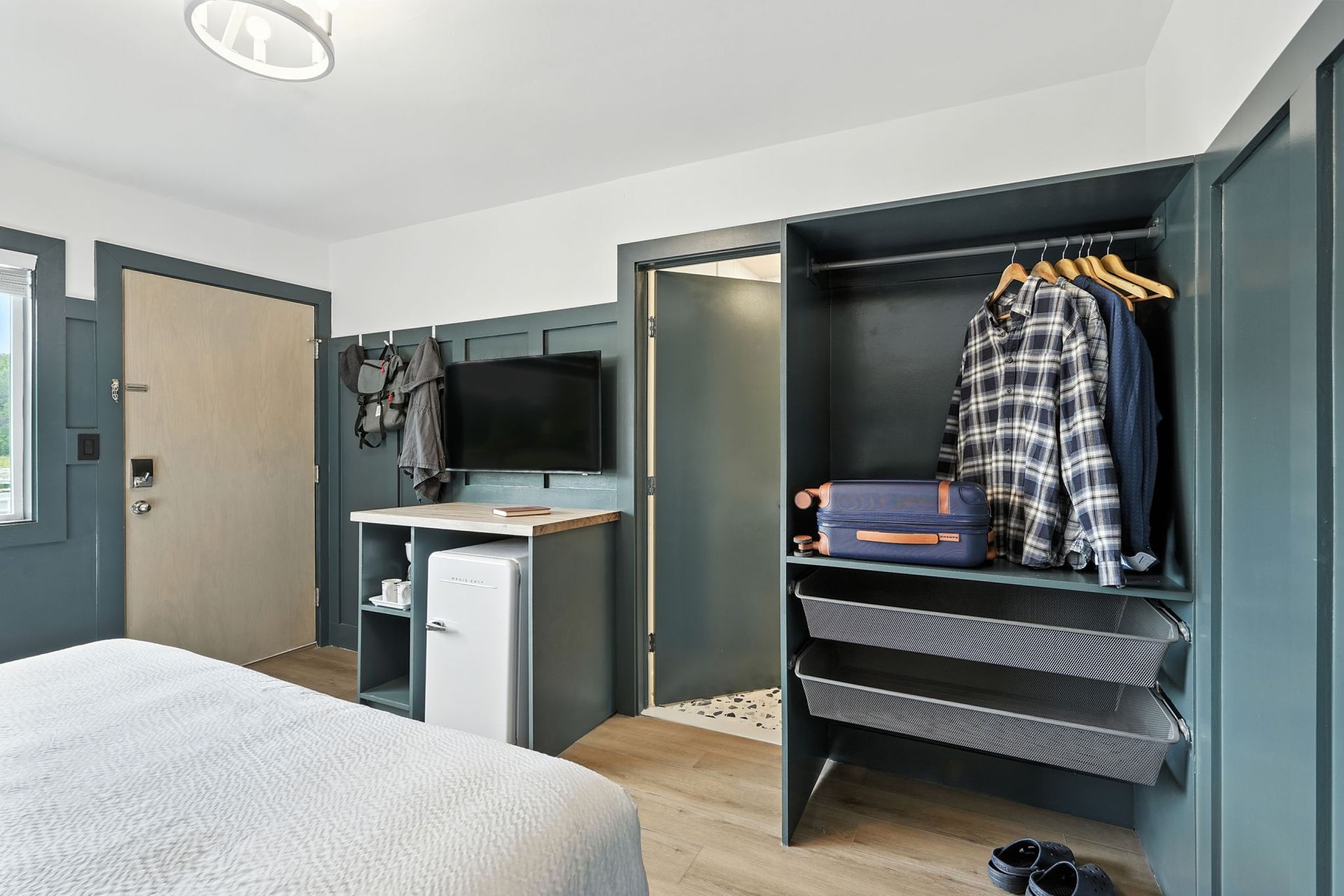 A hotel room interior features a bed, closet with clothes, TV, and small fridge.