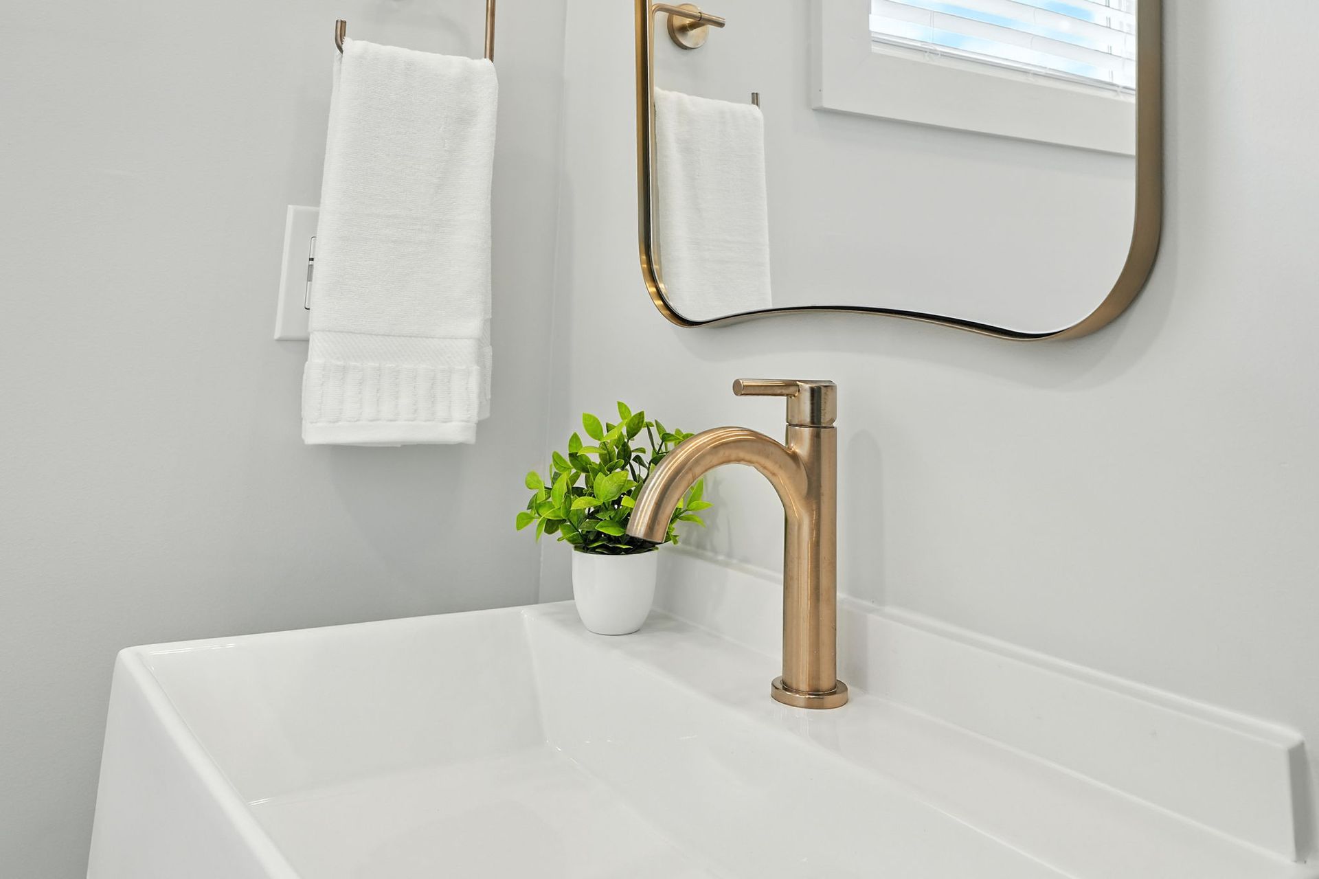 A modern bathroom with a white sink, gold faucet and mirror. A small plant sits next to the faucet.