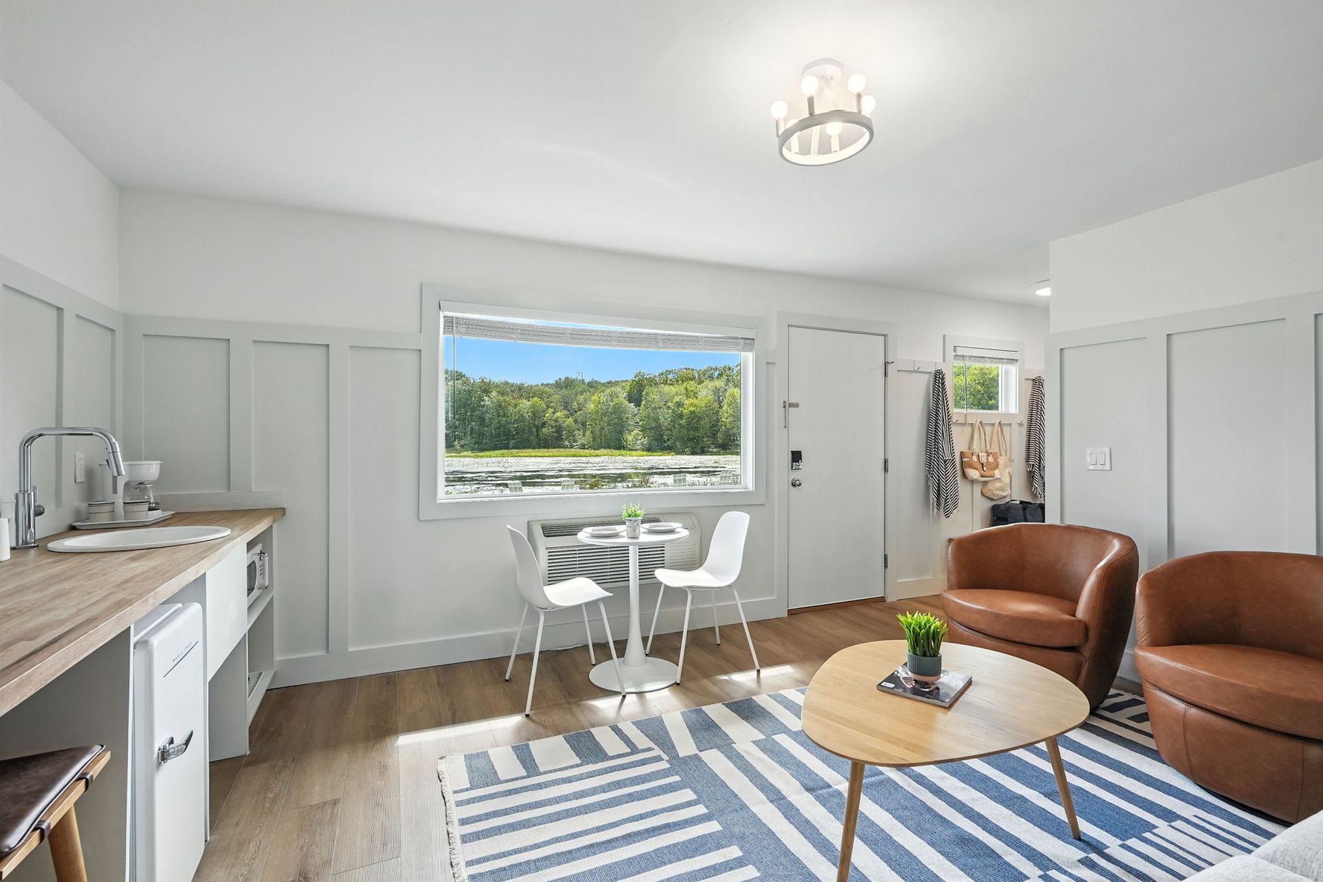 Small, bright studio apartment with kitchen, seating, and large window overlooking a natural landscape.