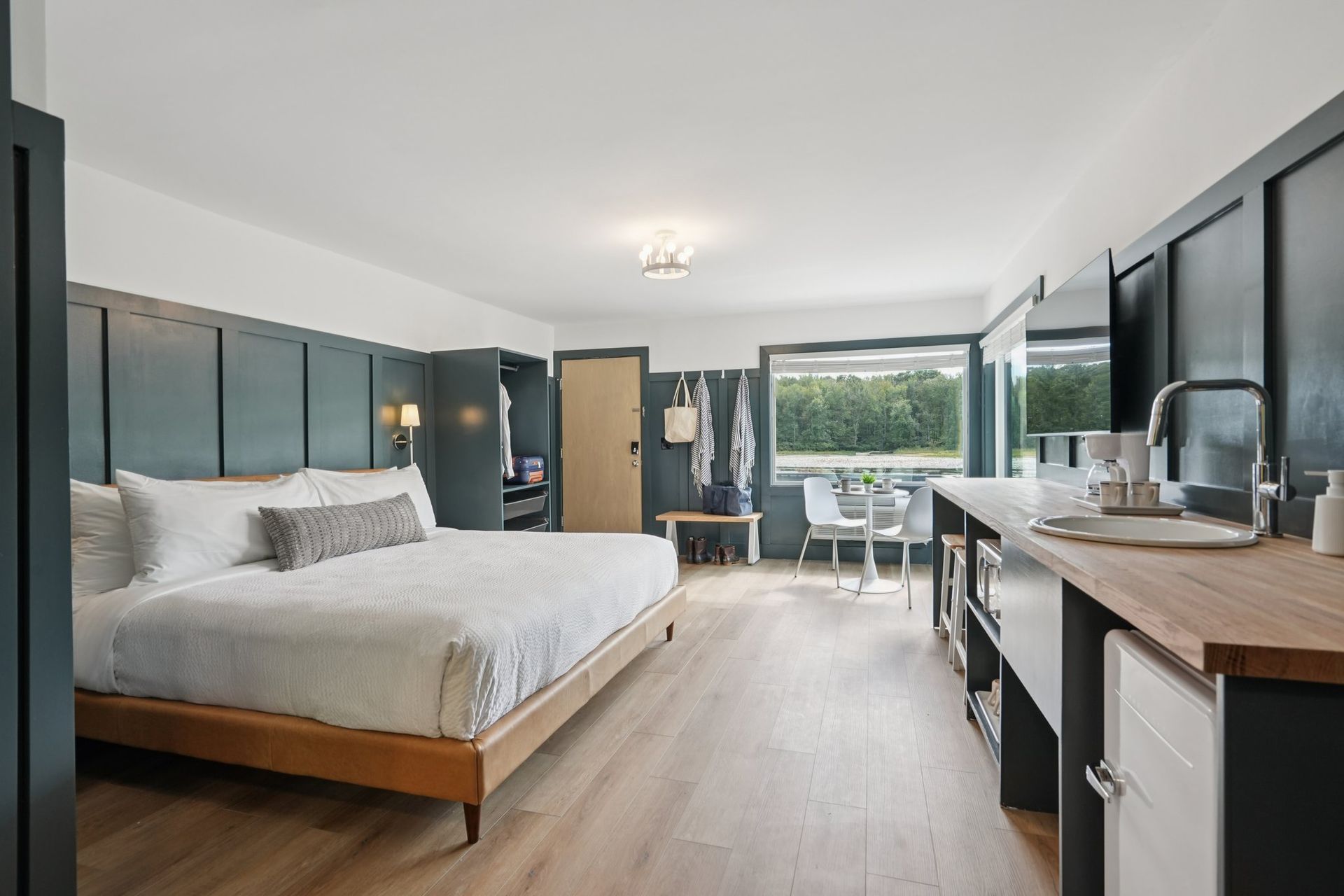 Hotel room with bed, kitchenette, and table overlooking outdoor area. Teal walls and wood accents.