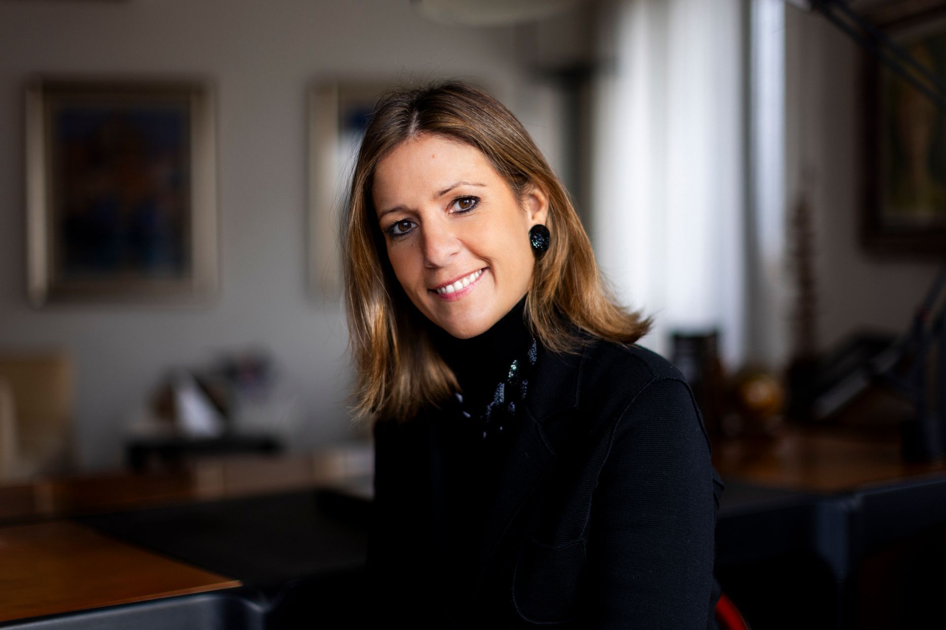 Woman in black turtleneck smiles, indoors with artwork in the background.