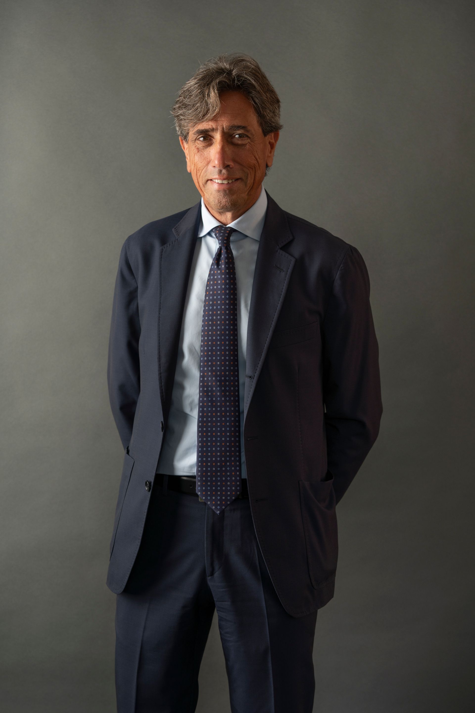 Man in a blue suit, hands behind his back, facing the camera against a gray background.