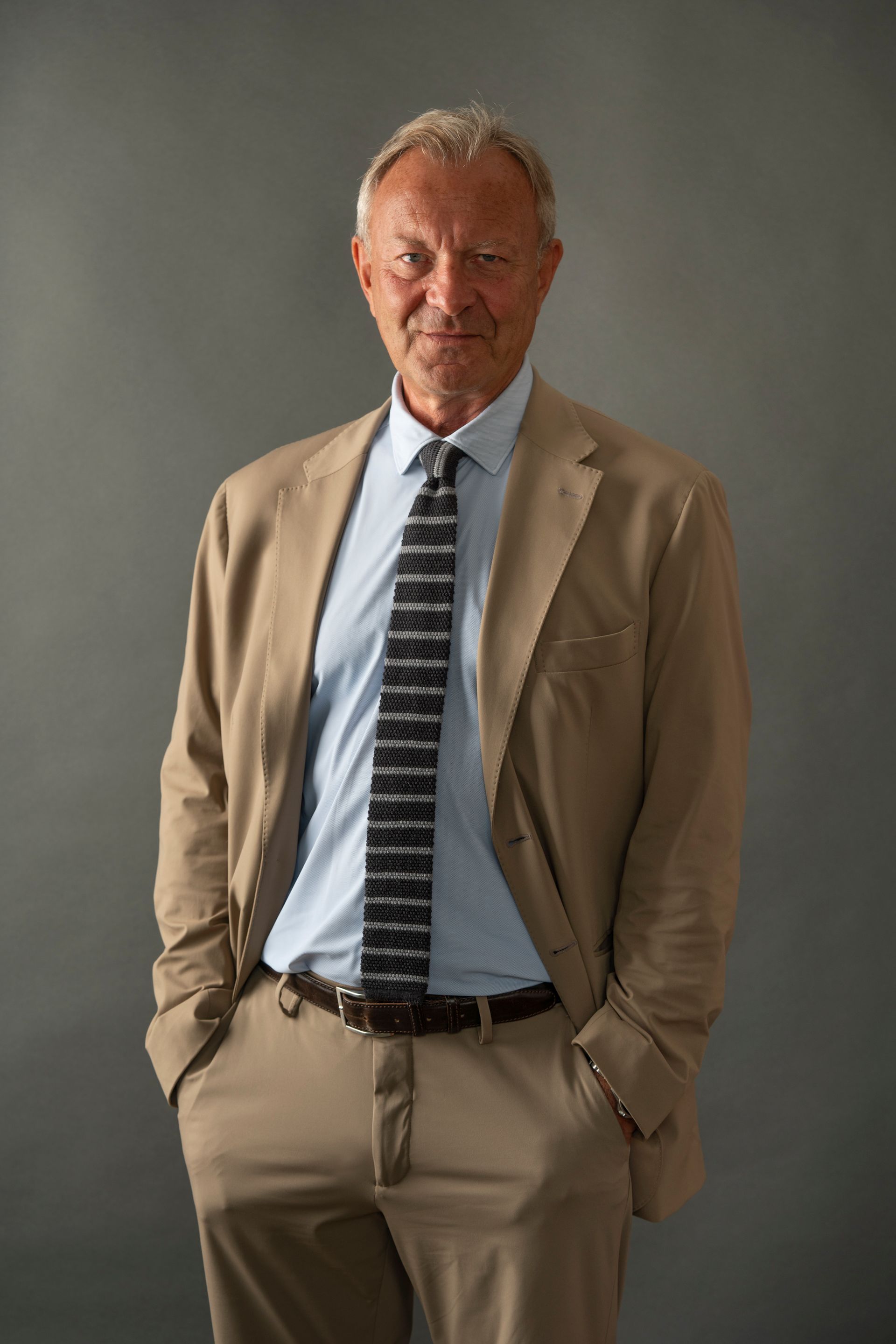 Man in tan suit, light blue shirt, patterned tie, hands in pockets; neutral background.