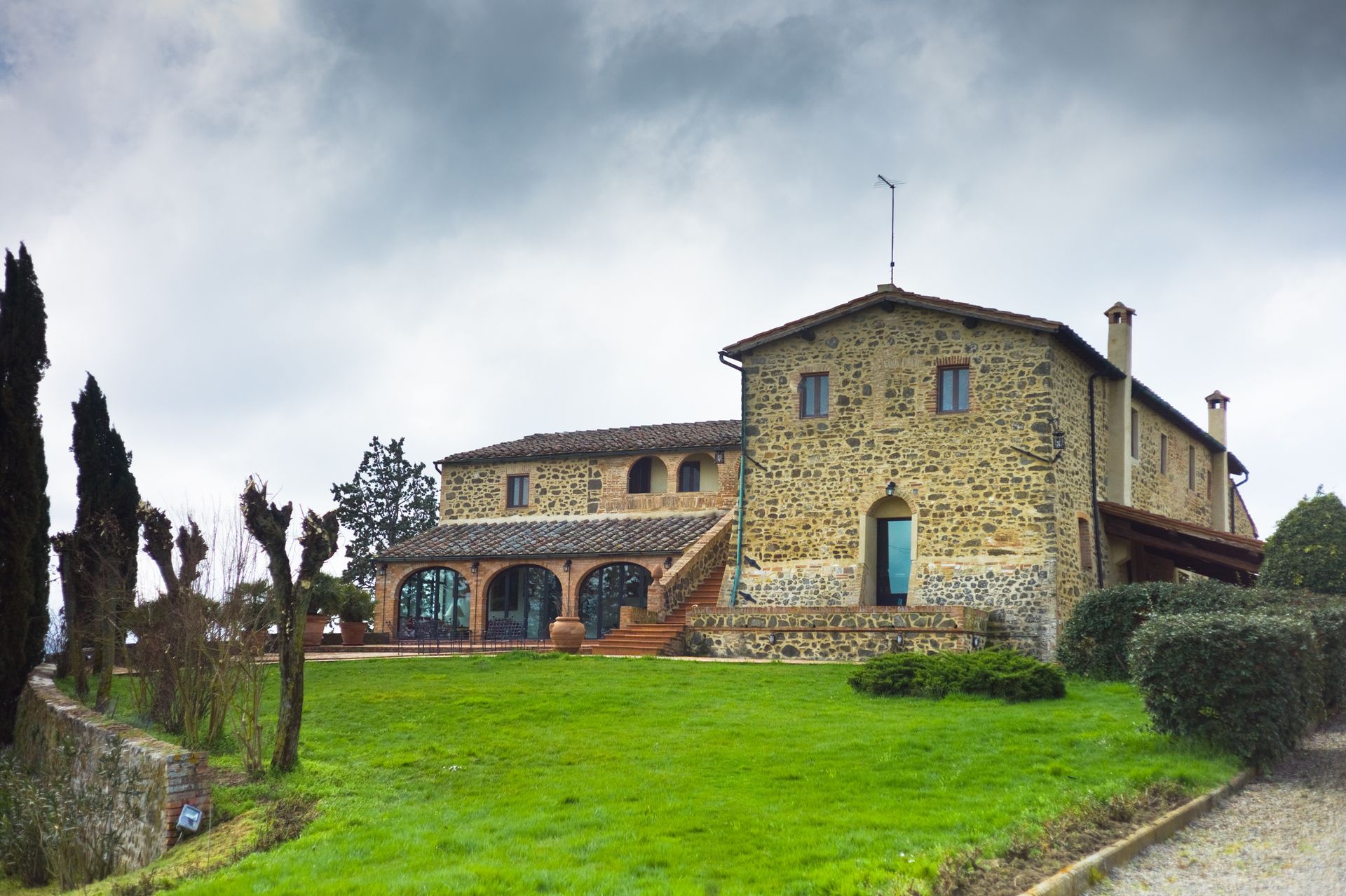 Stone building on a green lawn under a cloudy sky. Features a terrace and arched windows.