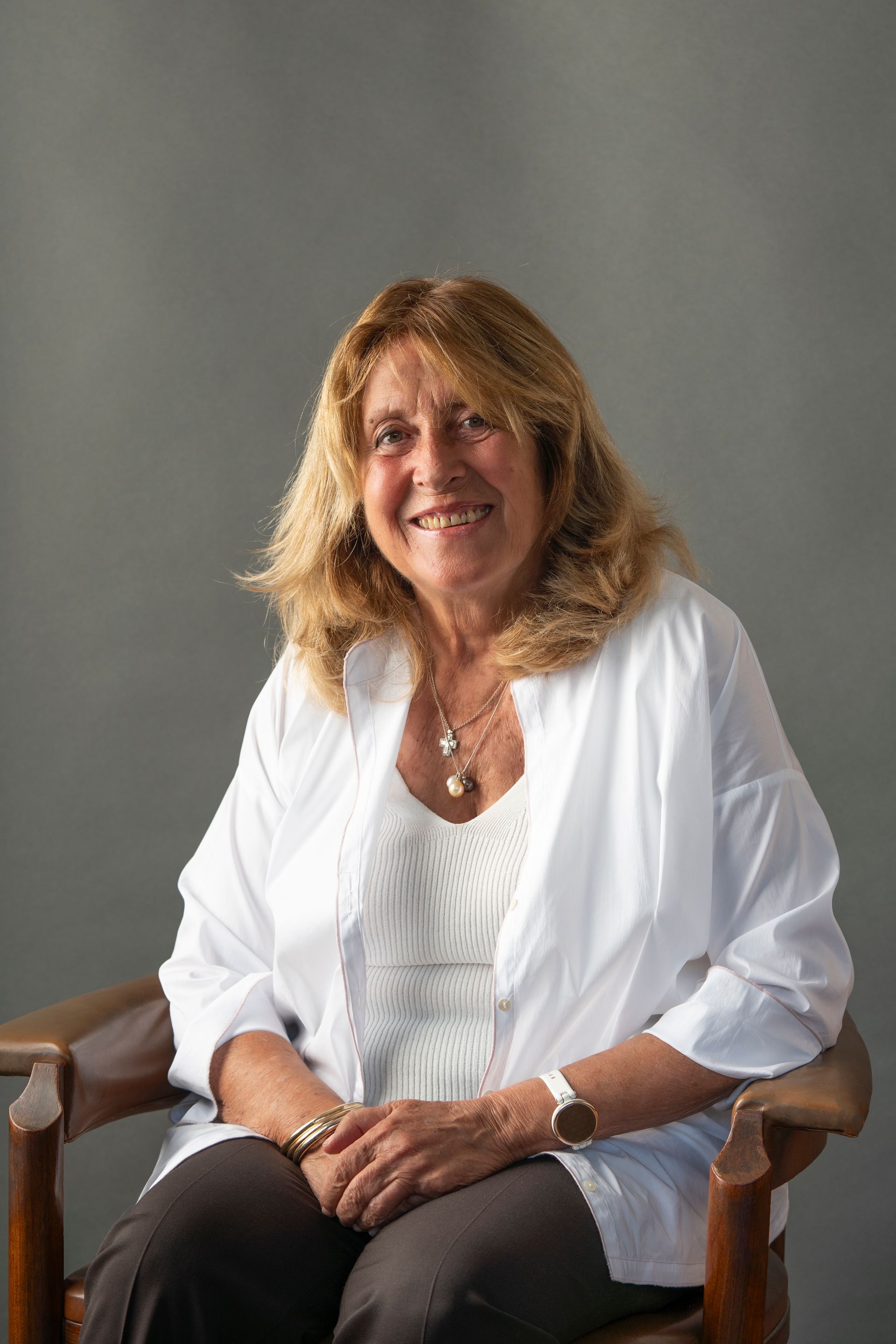 Woman with blonde hair, in a white shirt, sitting and smiling in front of a gray background.