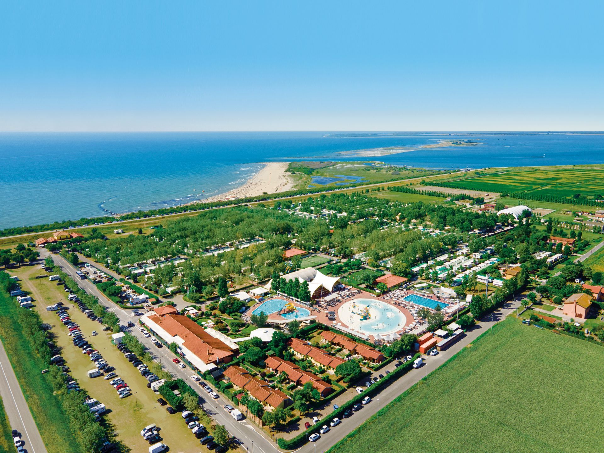 Vista aerea di una località balneare con piscine, edifici, alberi e una spiaggia, con cielo e mare azzurri.