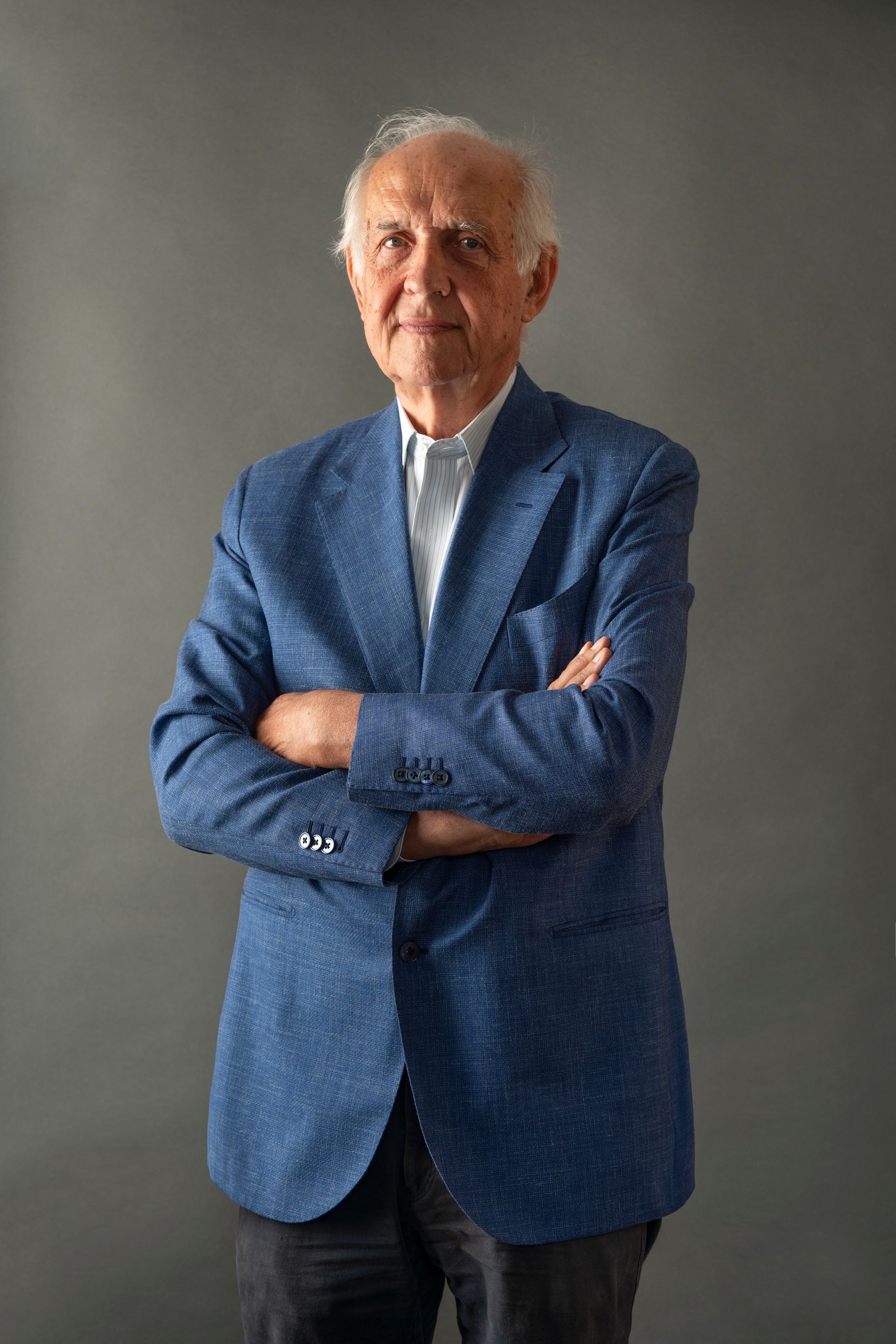 Man in blue blazer with arms crossed, standing against a gray backdrop.