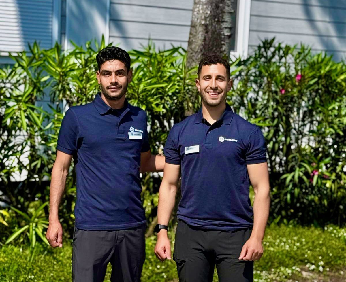 Due uomini in camicia blu, in piedi fuori, sorridenti.