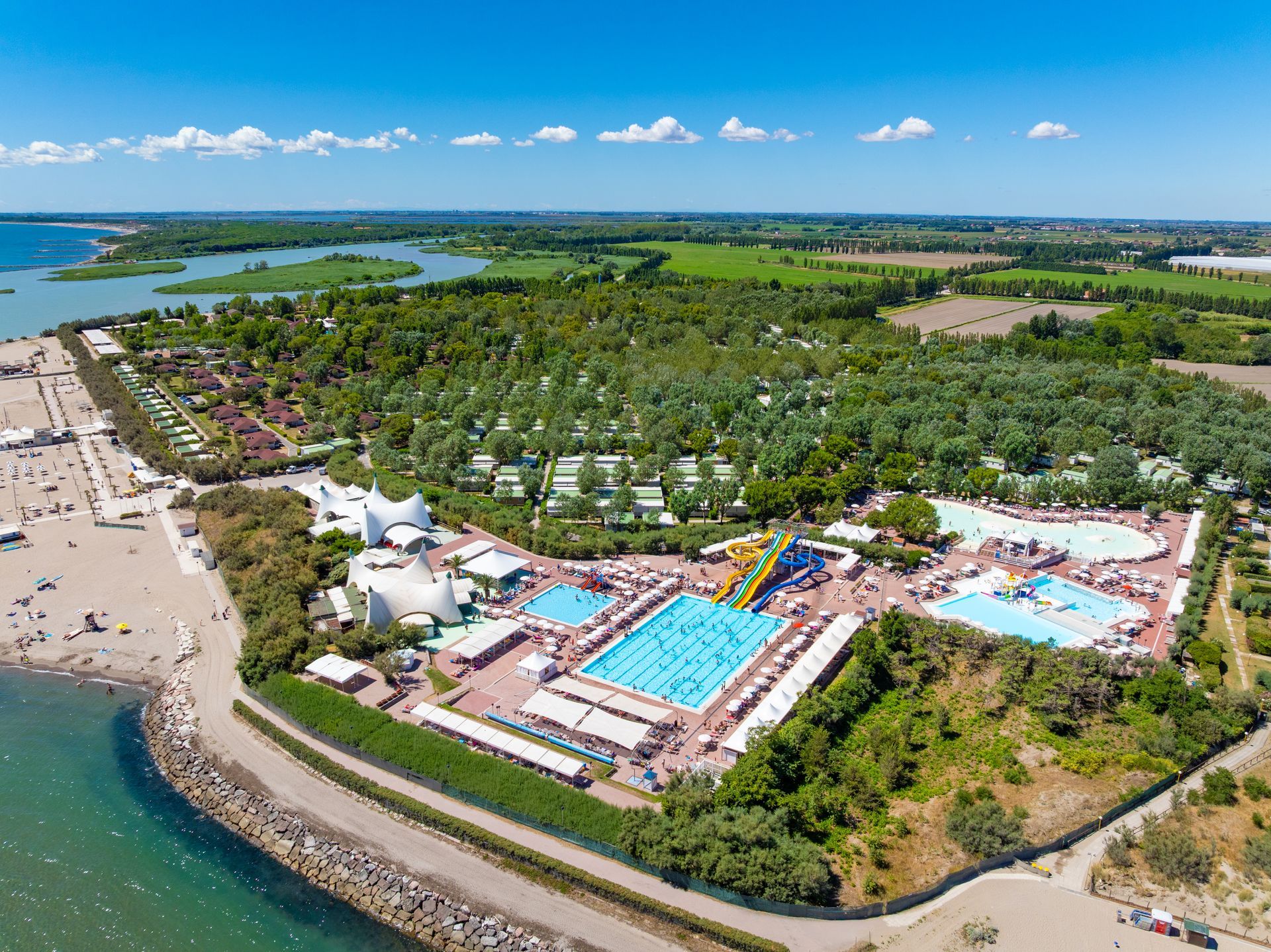Aerial view of a beach resort with pools, beach, and lush green trees under a bright blue sky.