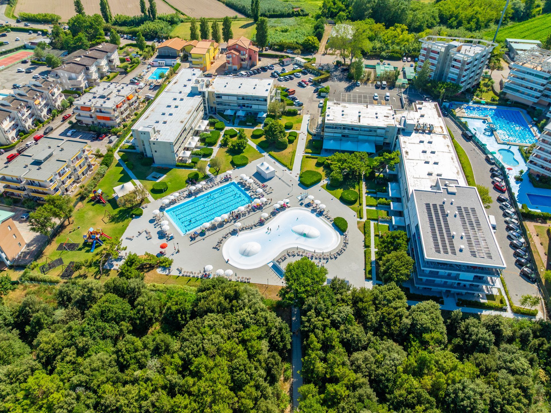 Aerial view of a resort with pools, buildings, trees, and a sunny sky.