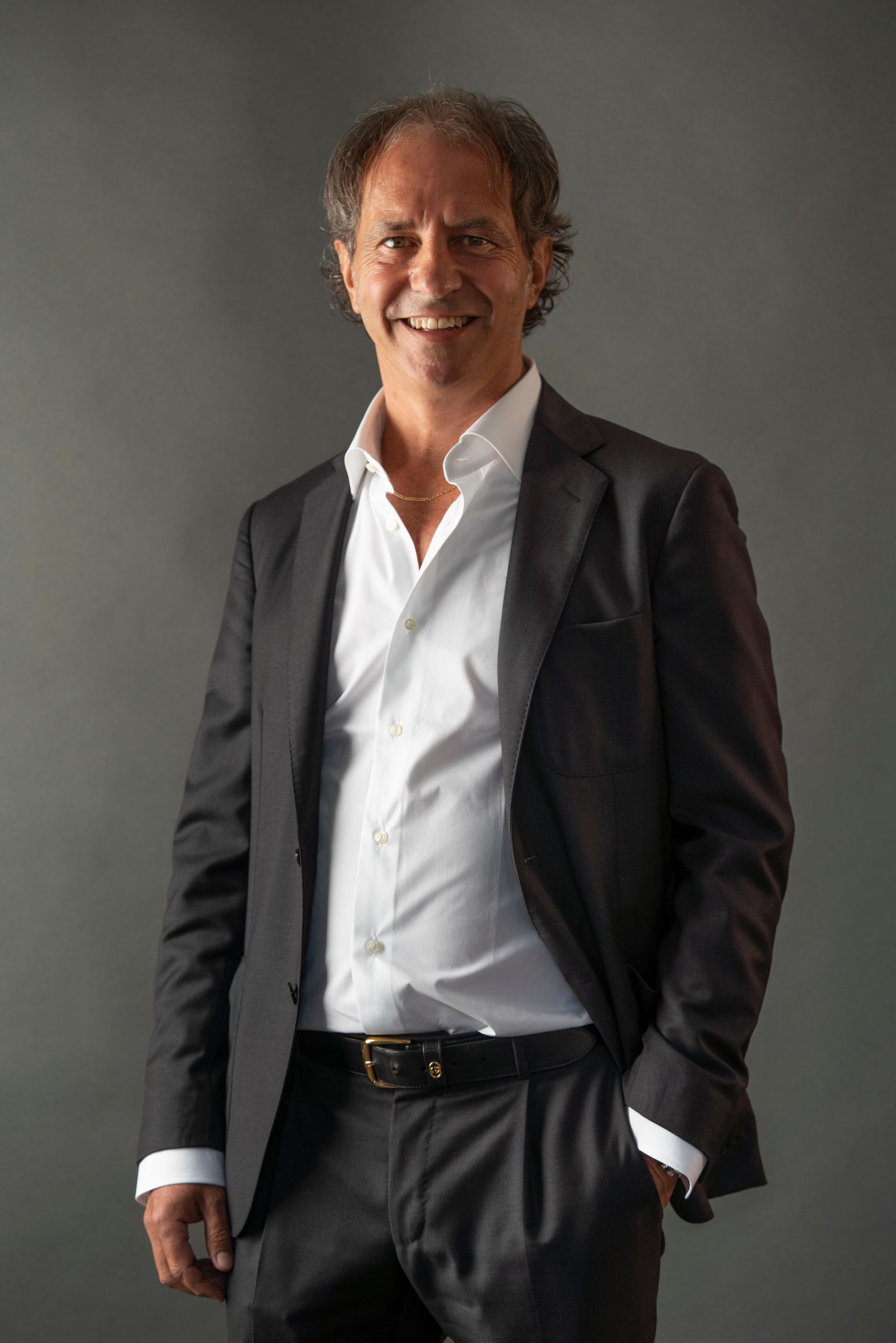 Man in a dark suit and open white shirt smiling, posing against a gray backdrop.
