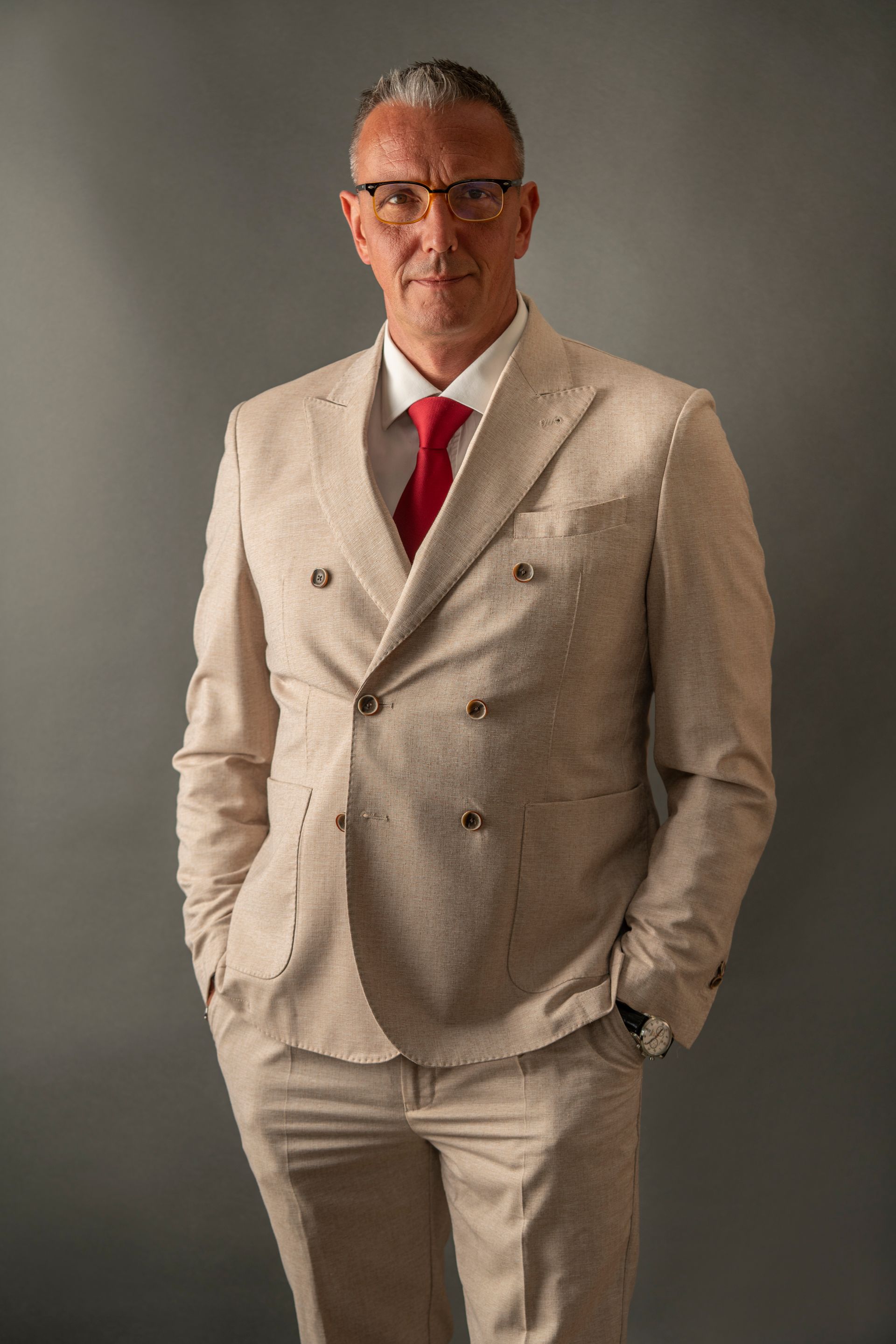 Man in beige suit with red tie, hands in pockets, standing in front of a gray background.