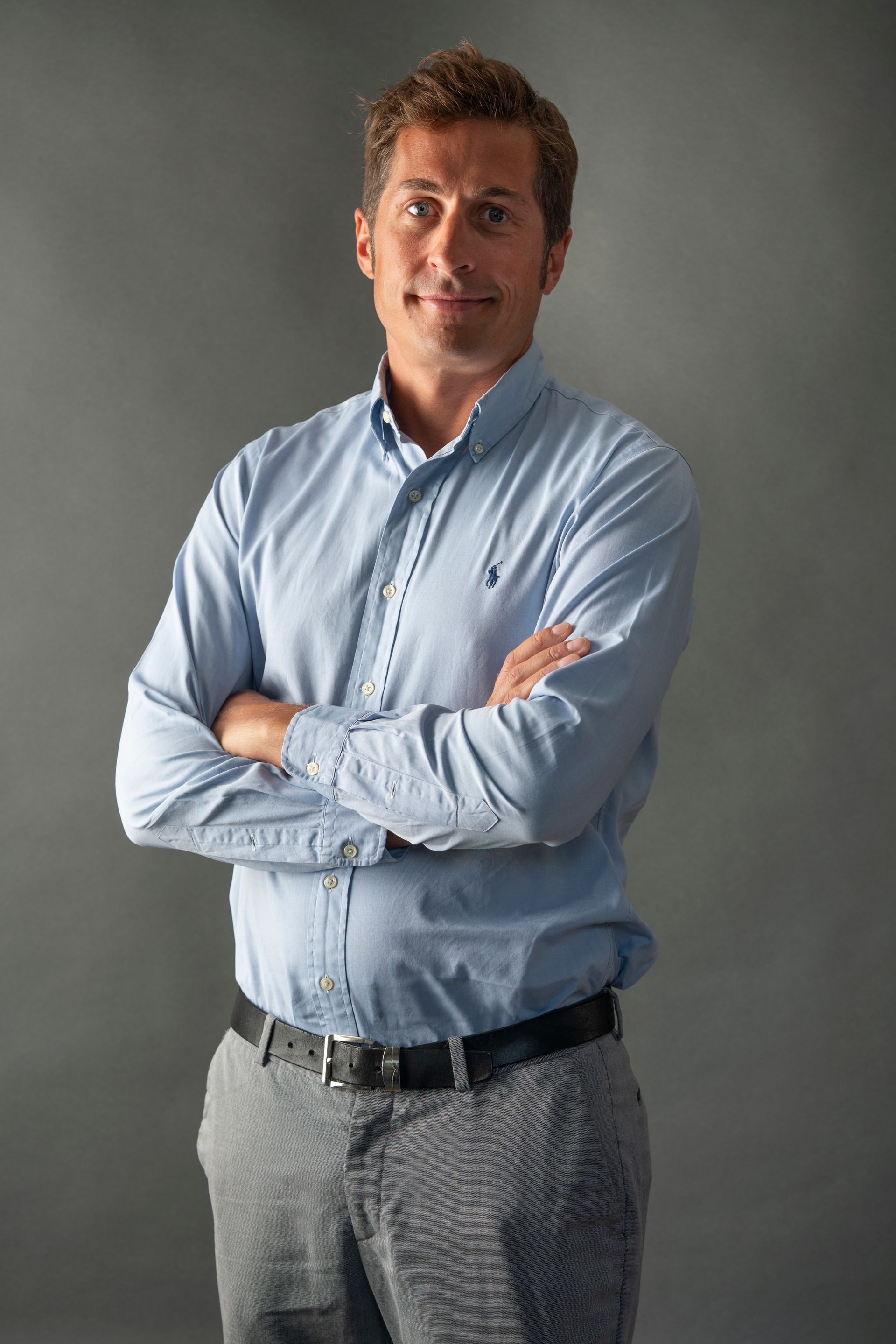 Man with crossed arms wearing a light blue shirt, gray pants, and a black belt, in a studio setting.