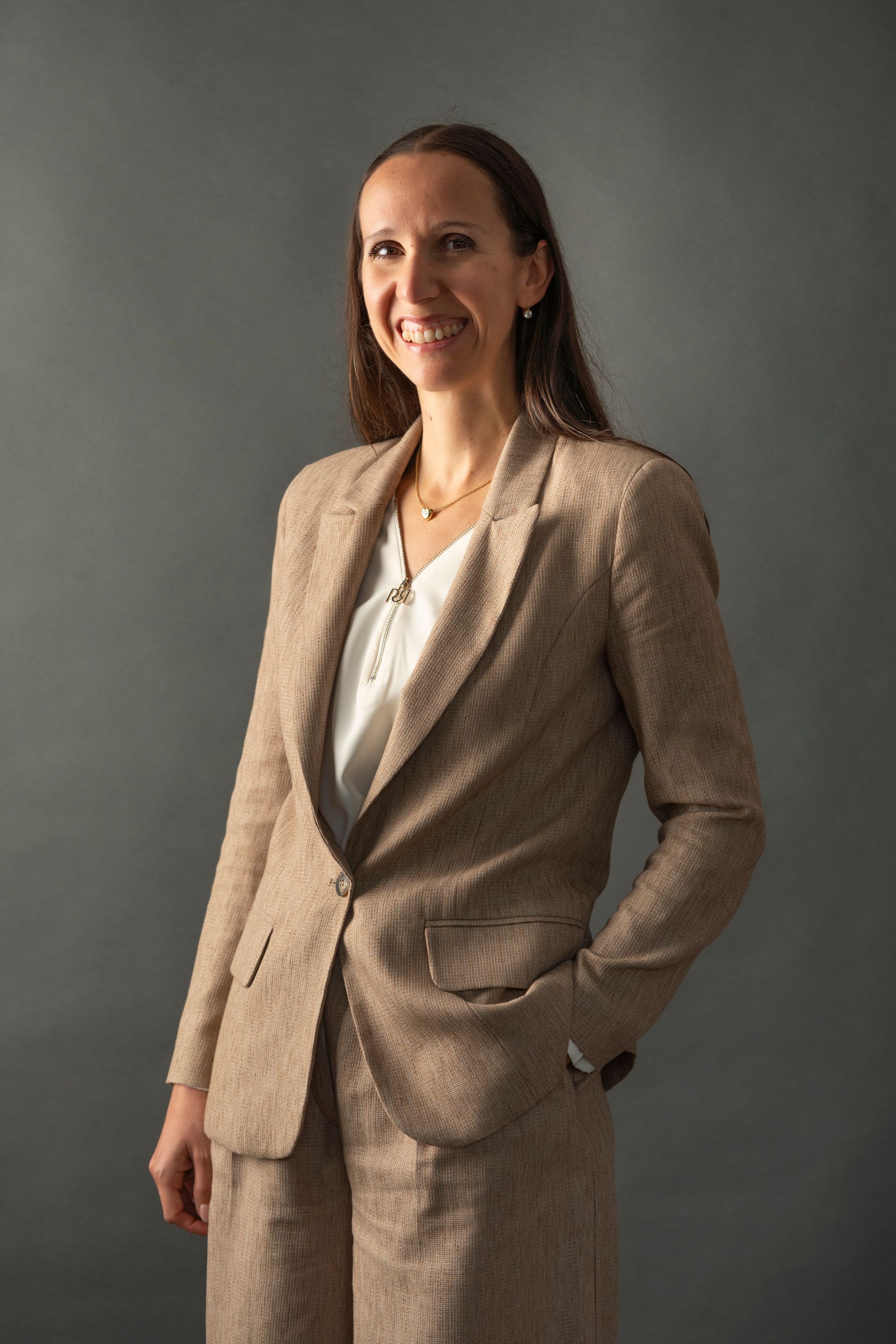 Donna in abito marrone chiaro, sorridente, mani in tasca, su uno sfondo grigio.