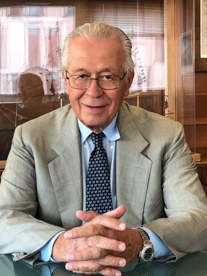 Man in glasses and suit, smiling with hands clasped, in an office setting.