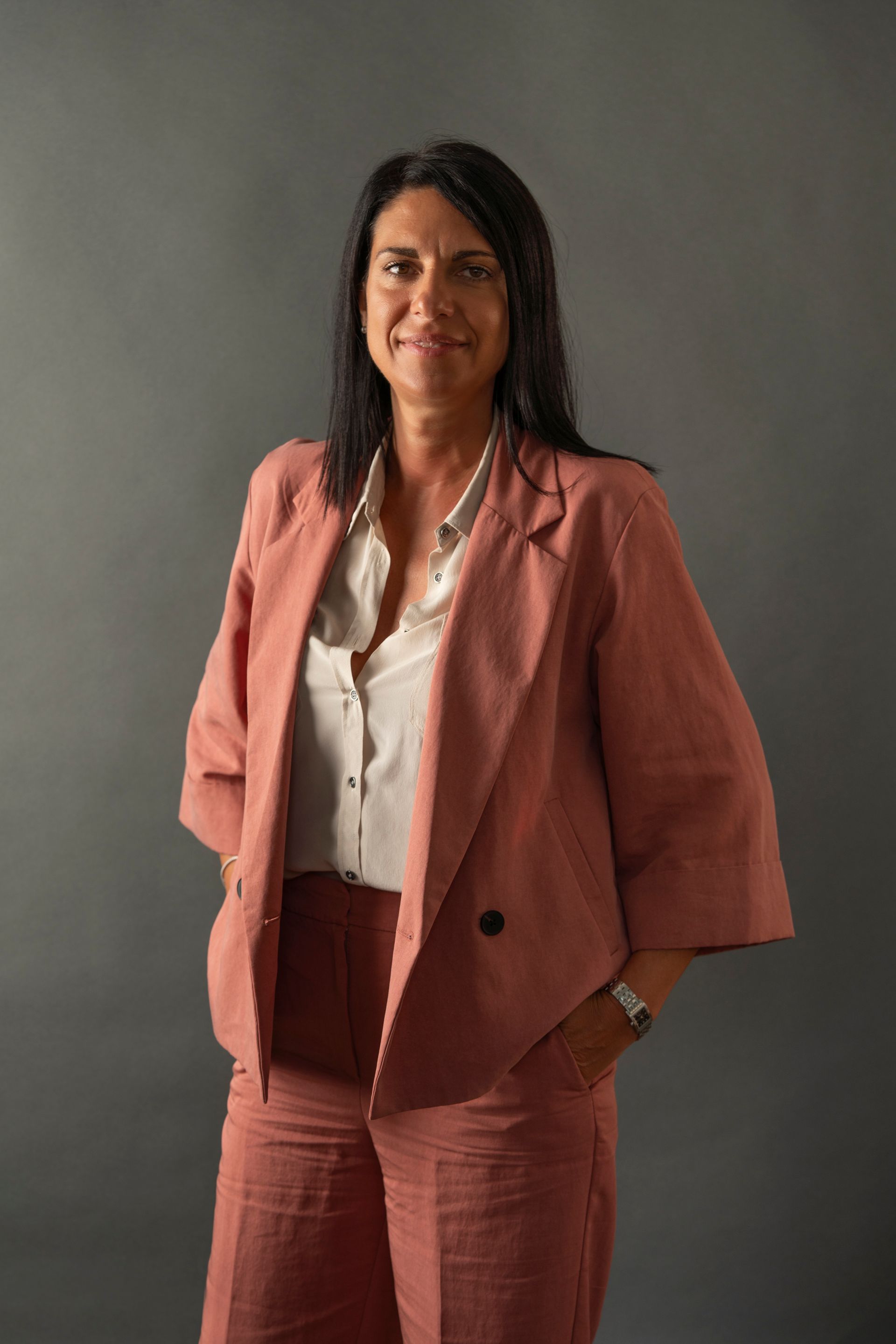 Woman with dark hair in a pink suit and white shirt, smiling, hands in pockets, against a gray backdrop.