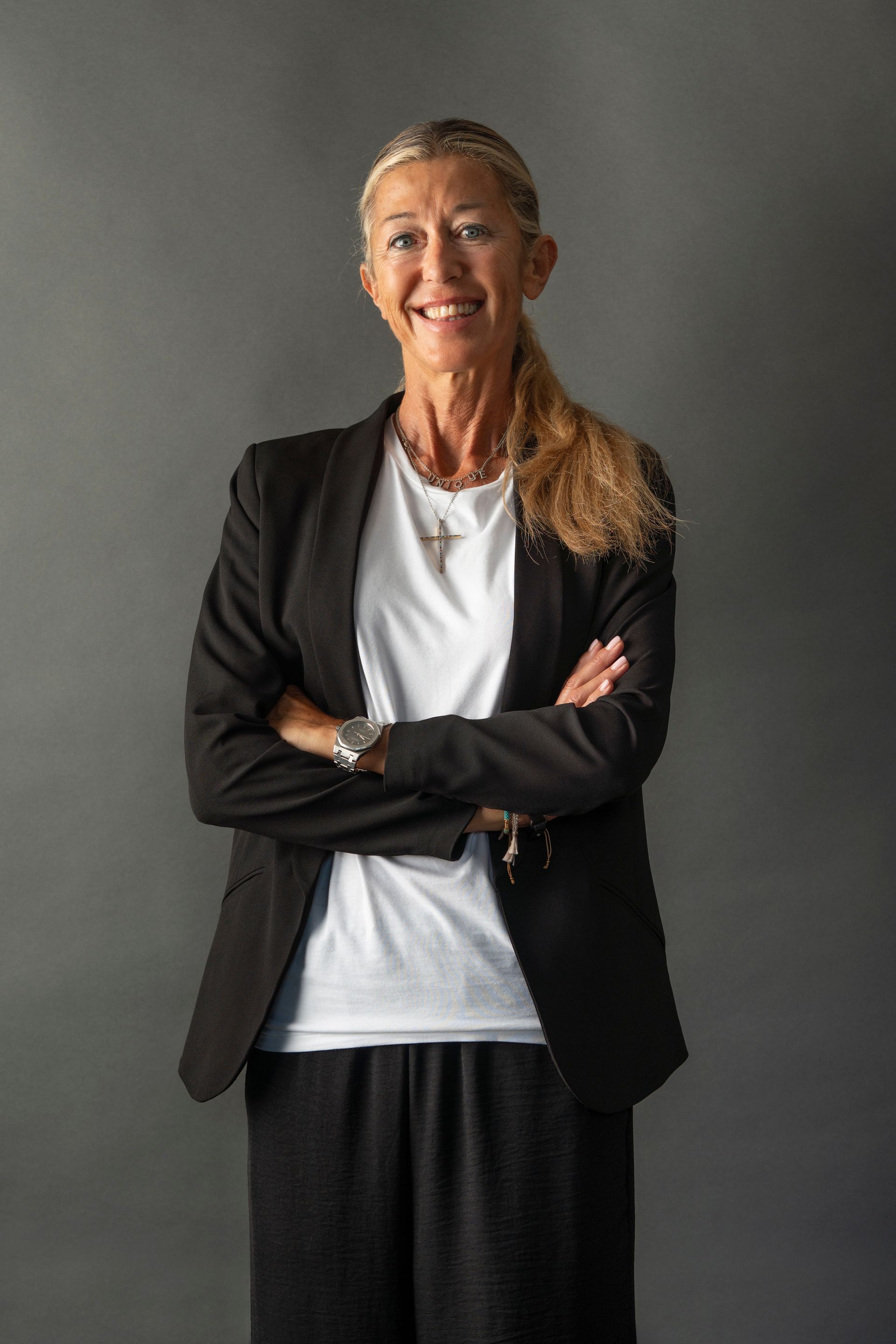 Woman in black blazer, arms crossed, smiling at the camera. Blonde hair, gray background.
