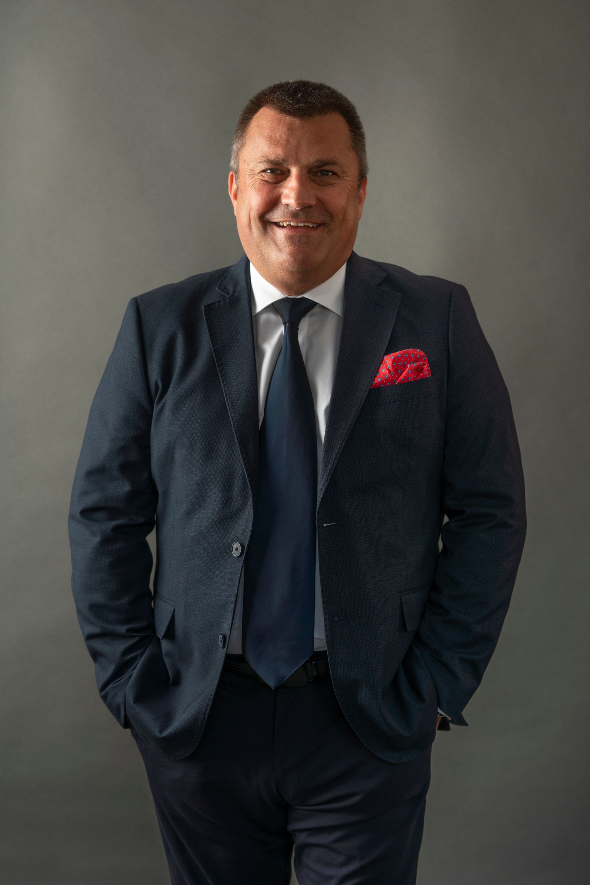 Man in a dark suit and tie, smiling, hands in pockets, with a red pocket square, against a gray backdrop.