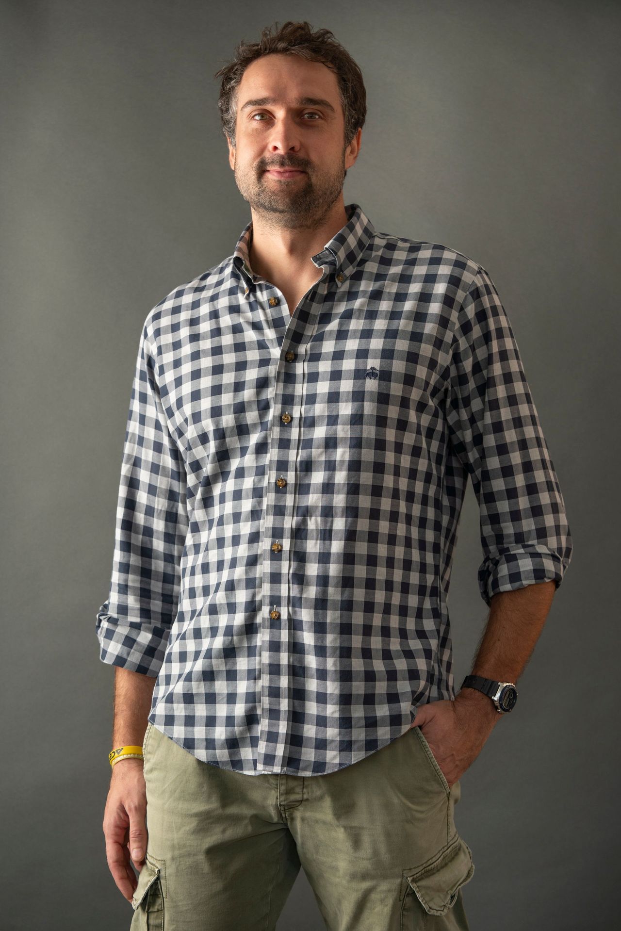 Man in plaid shirt and cargo pants, hand in pocket, smiling. Gray background.