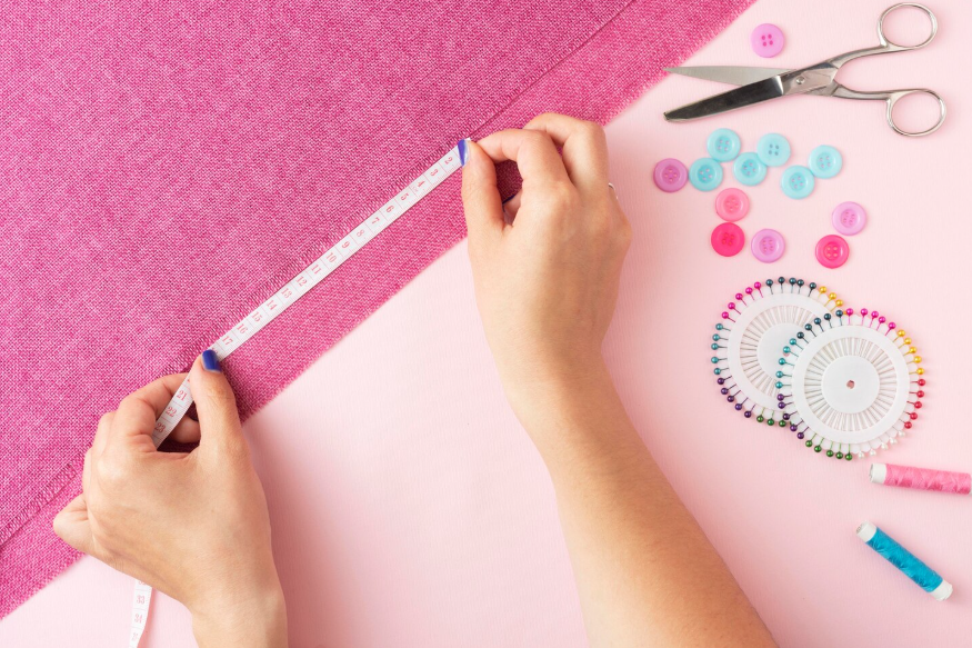 Manos midiendo tela rosa con una cinta métrica, herramientas de costura esparcidas cerca.