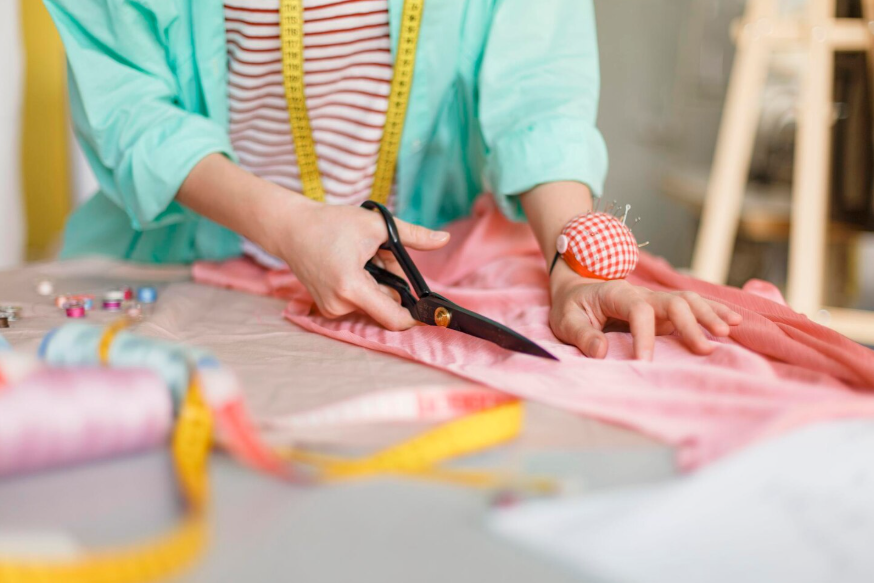 Persona cortando tela rosa con tijeras sobre una mesa; suministros de costura cerca.