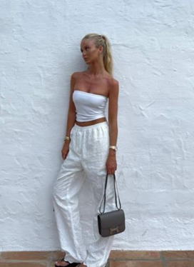 Una mujer vestida de blanco posa contra una pared blanca, sosteniendo un bolso.