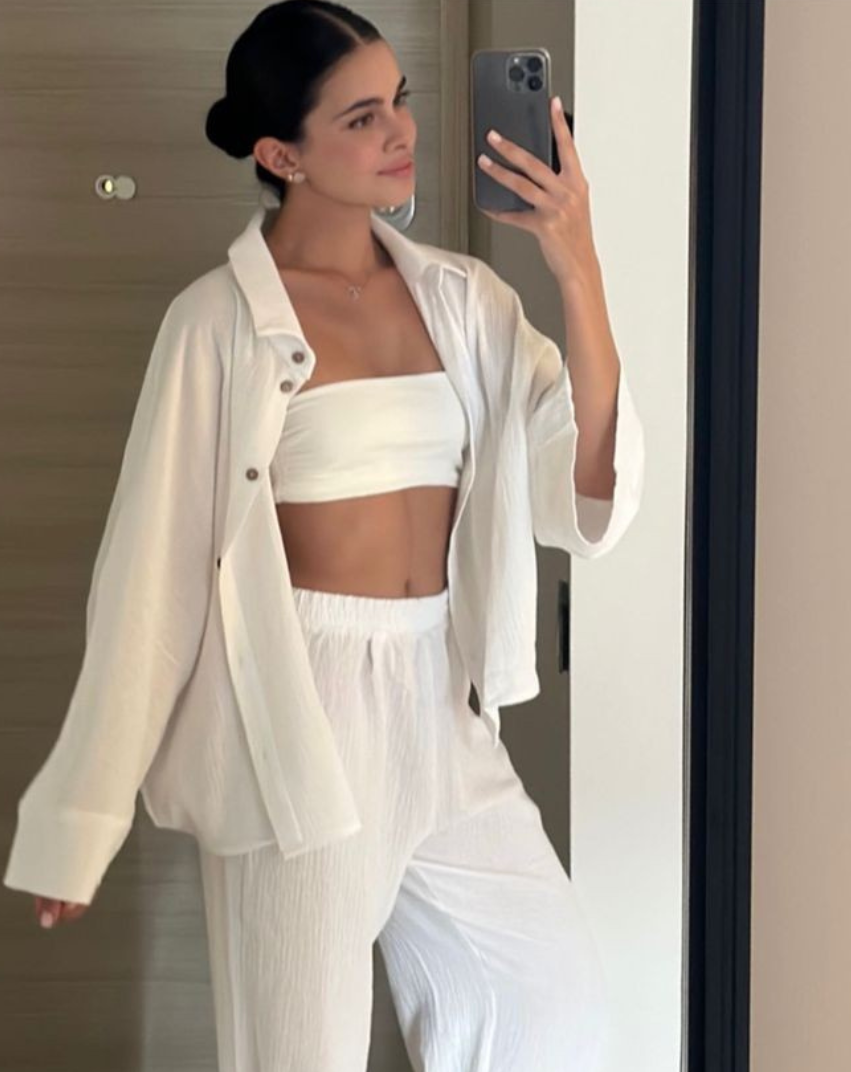 Mujer vestida de blanco tomándose una selfie frente al espejo. Lleva un top de tubo, una camisa abierta y pantalones