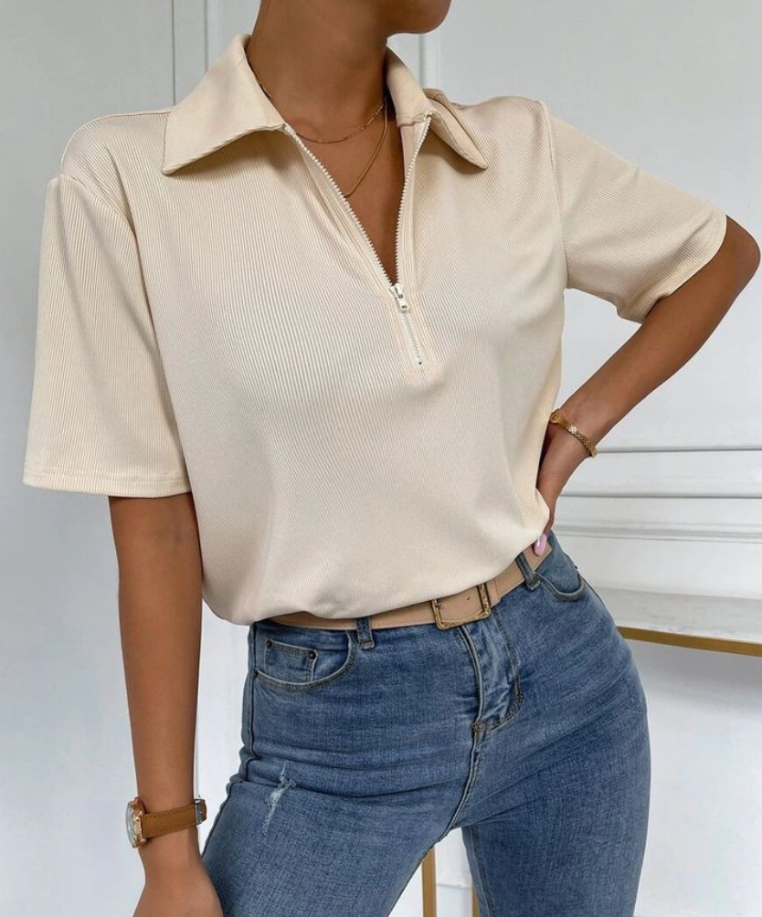 Mujer vestida con una blusa color crema con cremallera y pantalones vaqueros azules, de pie con la mano en la cadera.