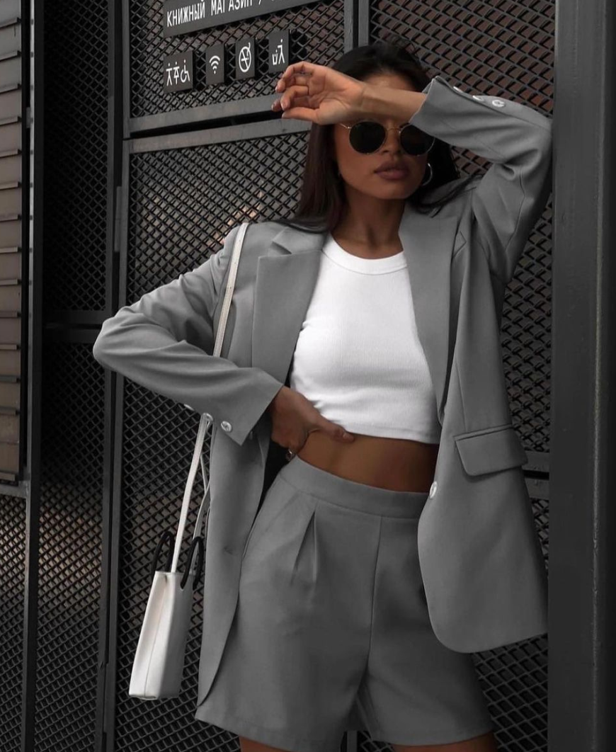 Mujer con blazer gris, pantalones cortos y top blanco, gafas de sol, mano en la frente, apoyada en una puerta de metal.