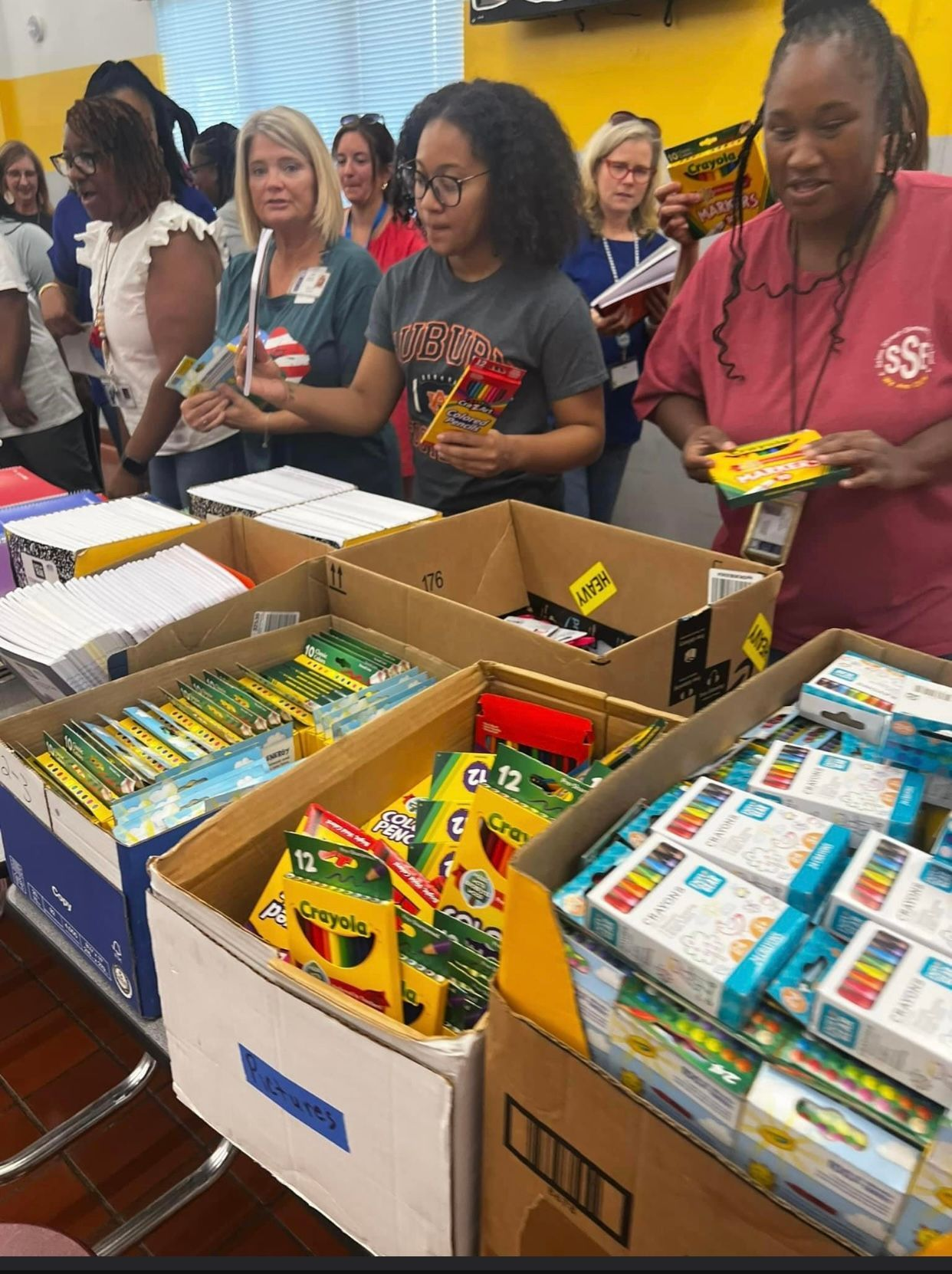 A group of people are standing around a table filled with boxes of crayons.