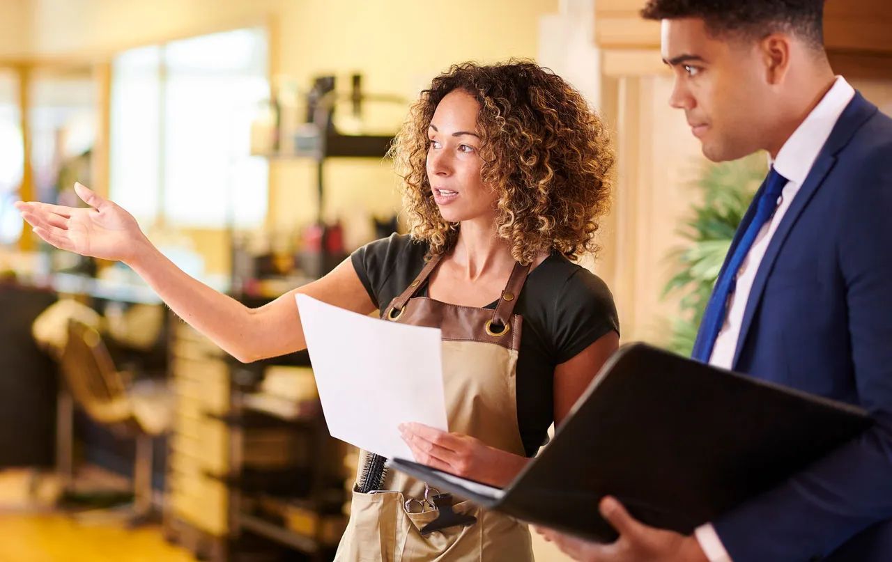 A man in a suit is standing next to a woman in an apron holding a clipboard.