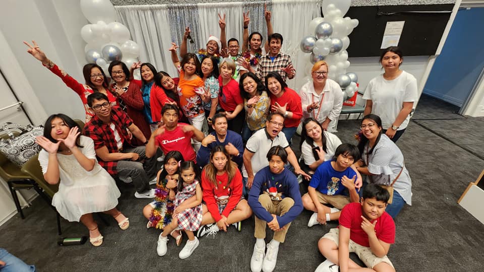Group photo: Diverse people, some children, indoors, some smiling, others making faces. Balloons in background.