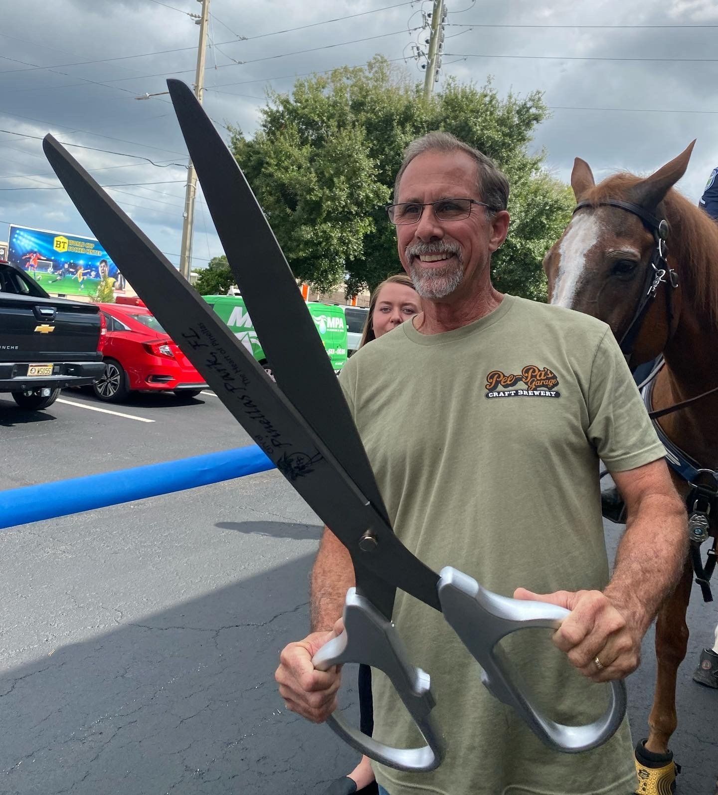 A man holding a pair of giant scissors with a horse in the background