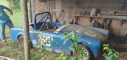 A man is standing next to a blue car parked in front of a chicken coop.