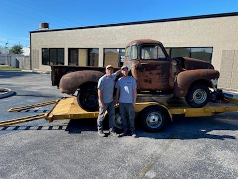 Two men are standing next to an old rusty truck on a trailer.