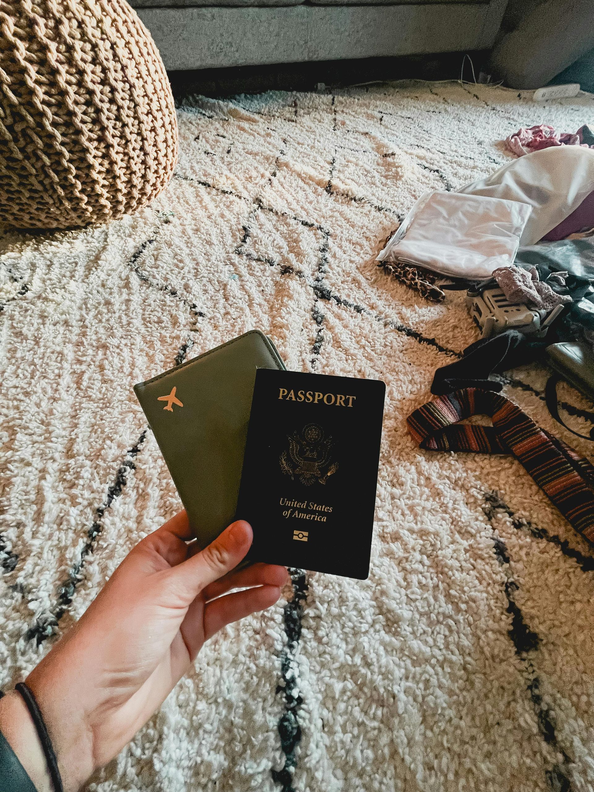 Hand holding a green passport holder and a black U.S. passport. The background is a patterned rug and a woven basket.