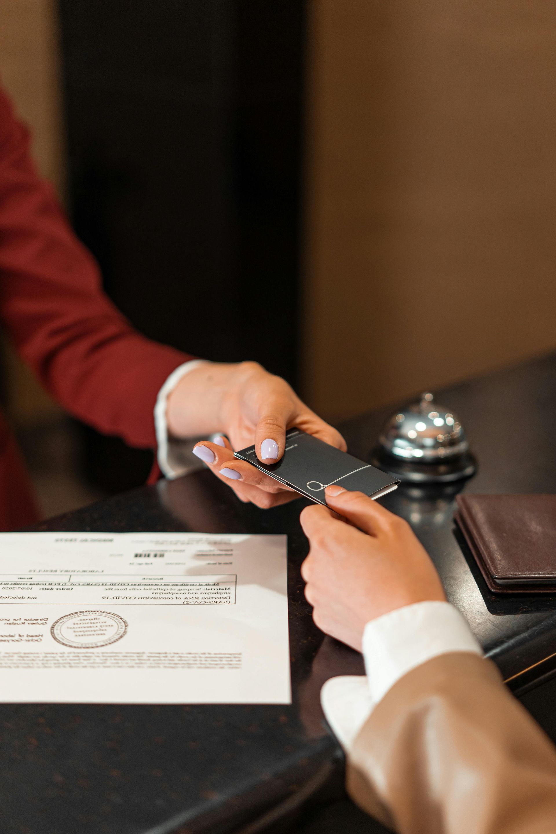 Person in a burgundy suit hands a black card to another person at a hotel reception desk, next to a bell and paperwork.