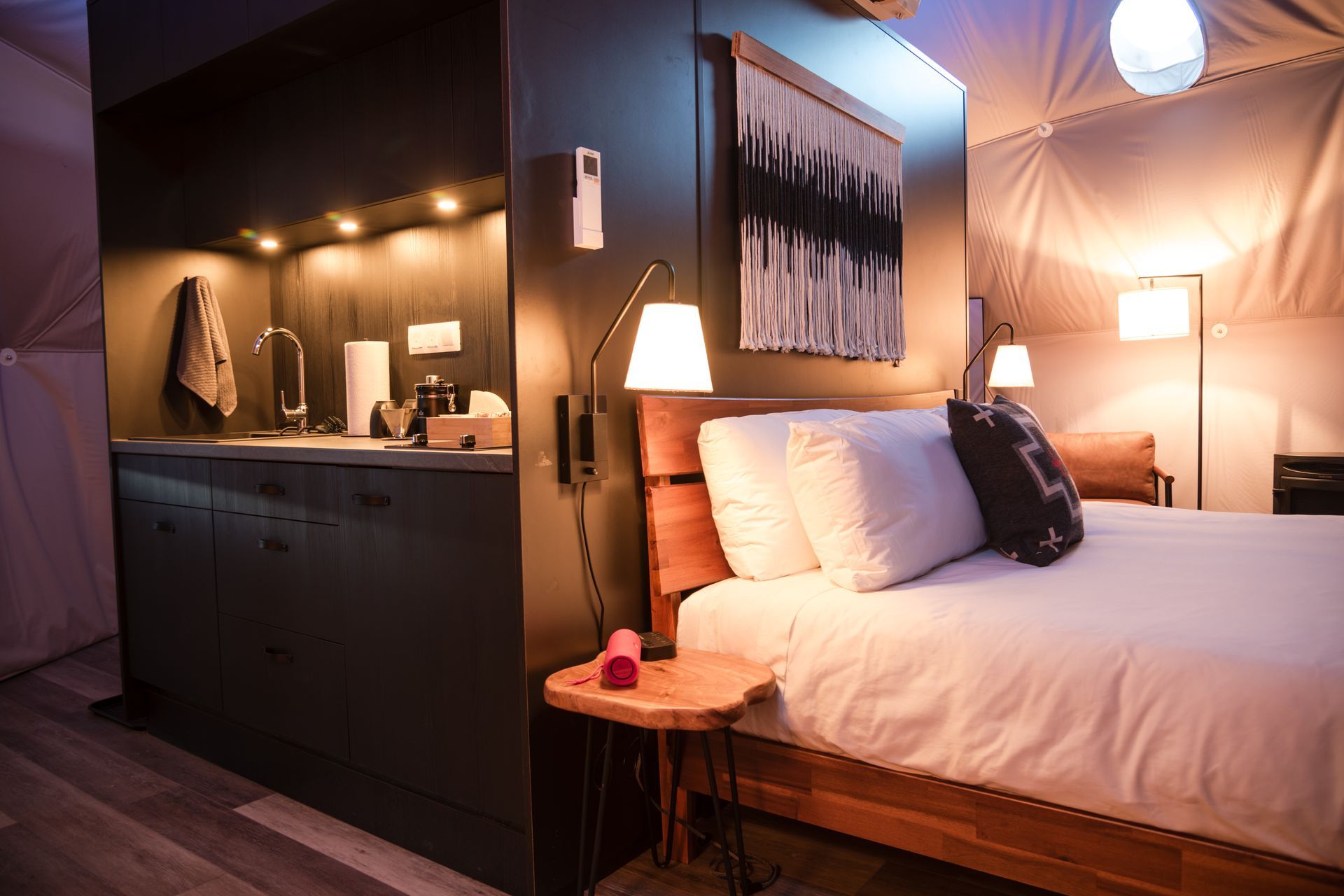 Bedroom with a bed, kitchenette, and artwork. Warm lighting and neutral colors create a cozy atmosphere.
