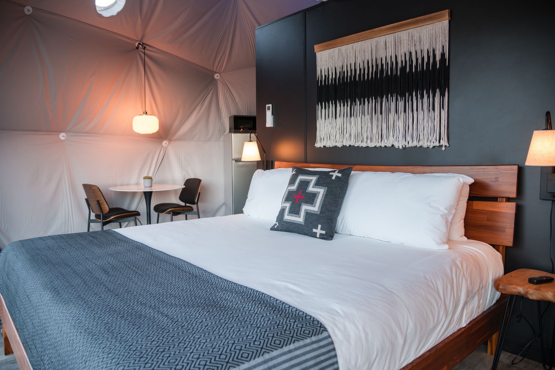 Bedroom inside a white dome. Bed with blue blanket, black wall, macrame art, small table with chairs.