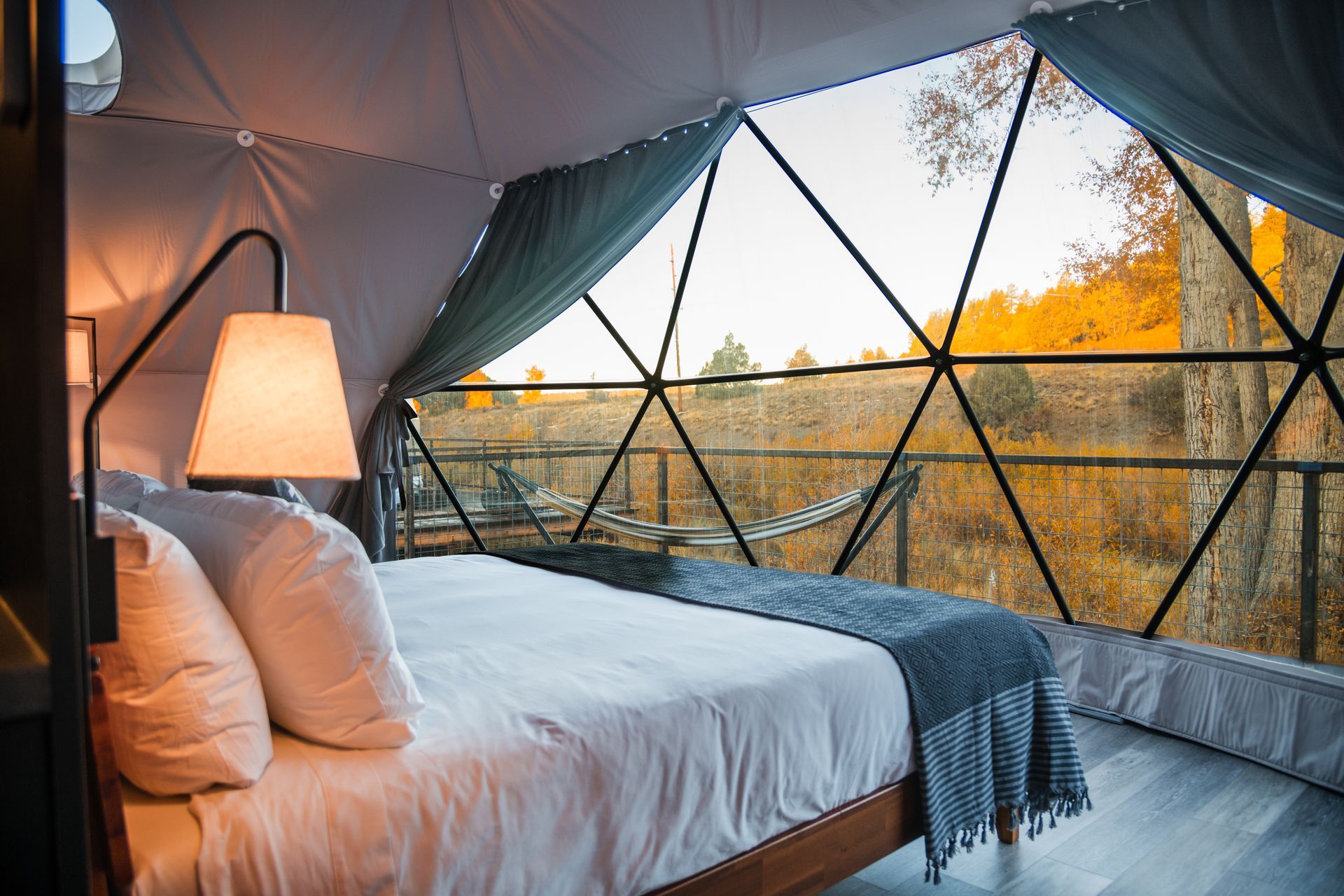 Cozy glamping dome interior with a bed, lamp, and view of fall foliage.