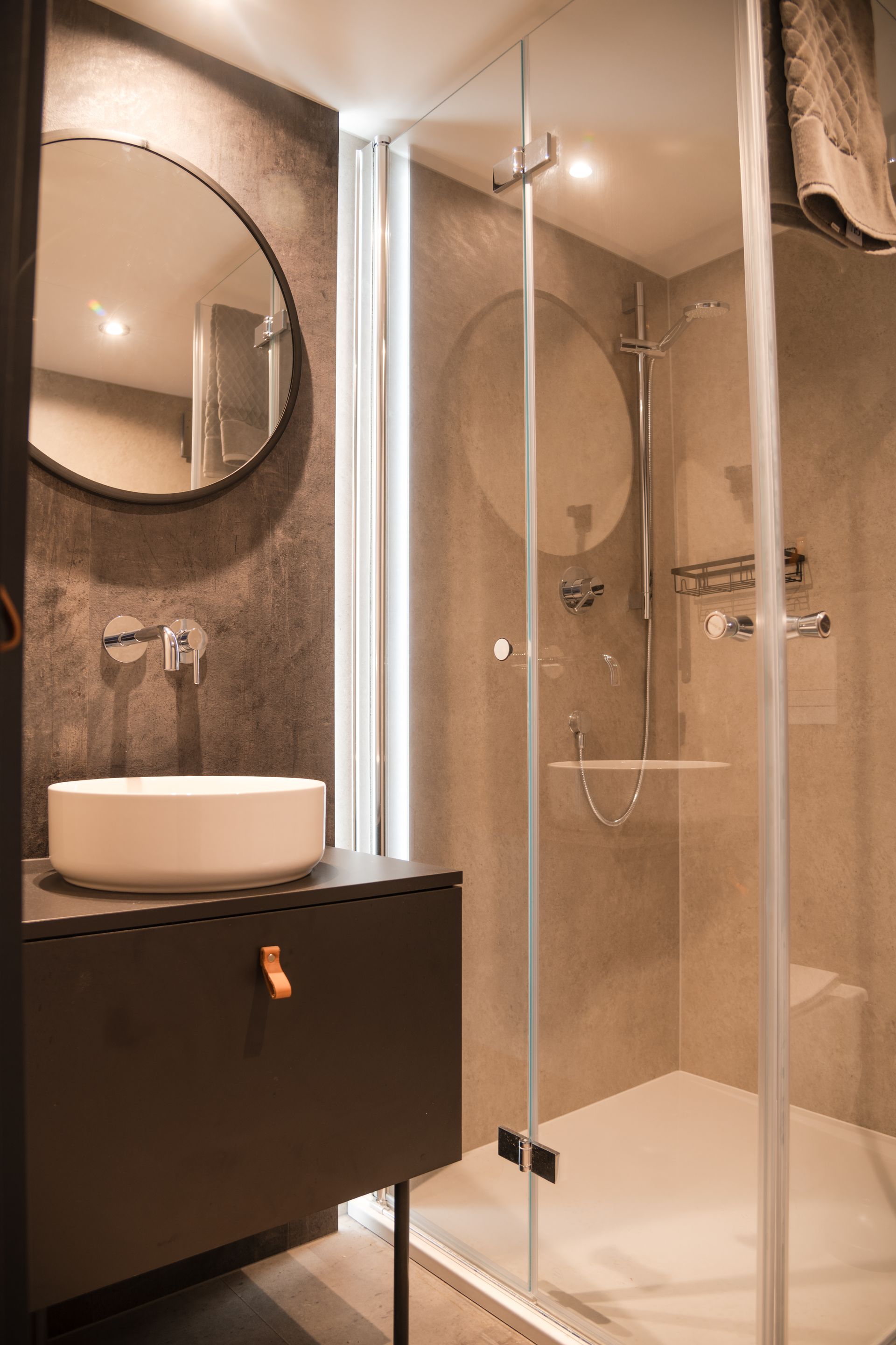 Modern bathroom with dark gray vanity, round mirror, glass shower, and neutral tile.
