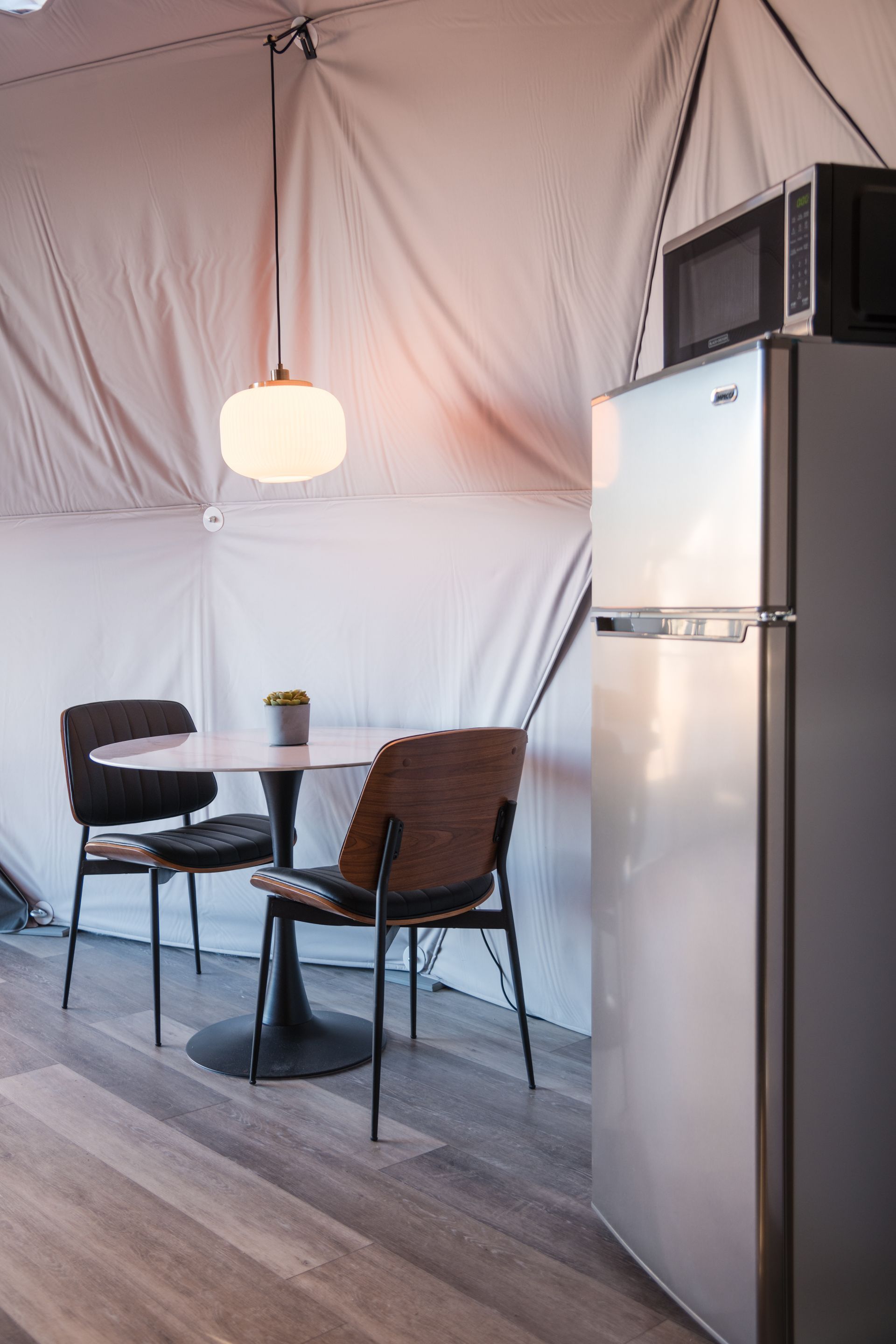 Interior of a glamping tent: small dining table with chairs, refrigerator, microwave, and pendant light.