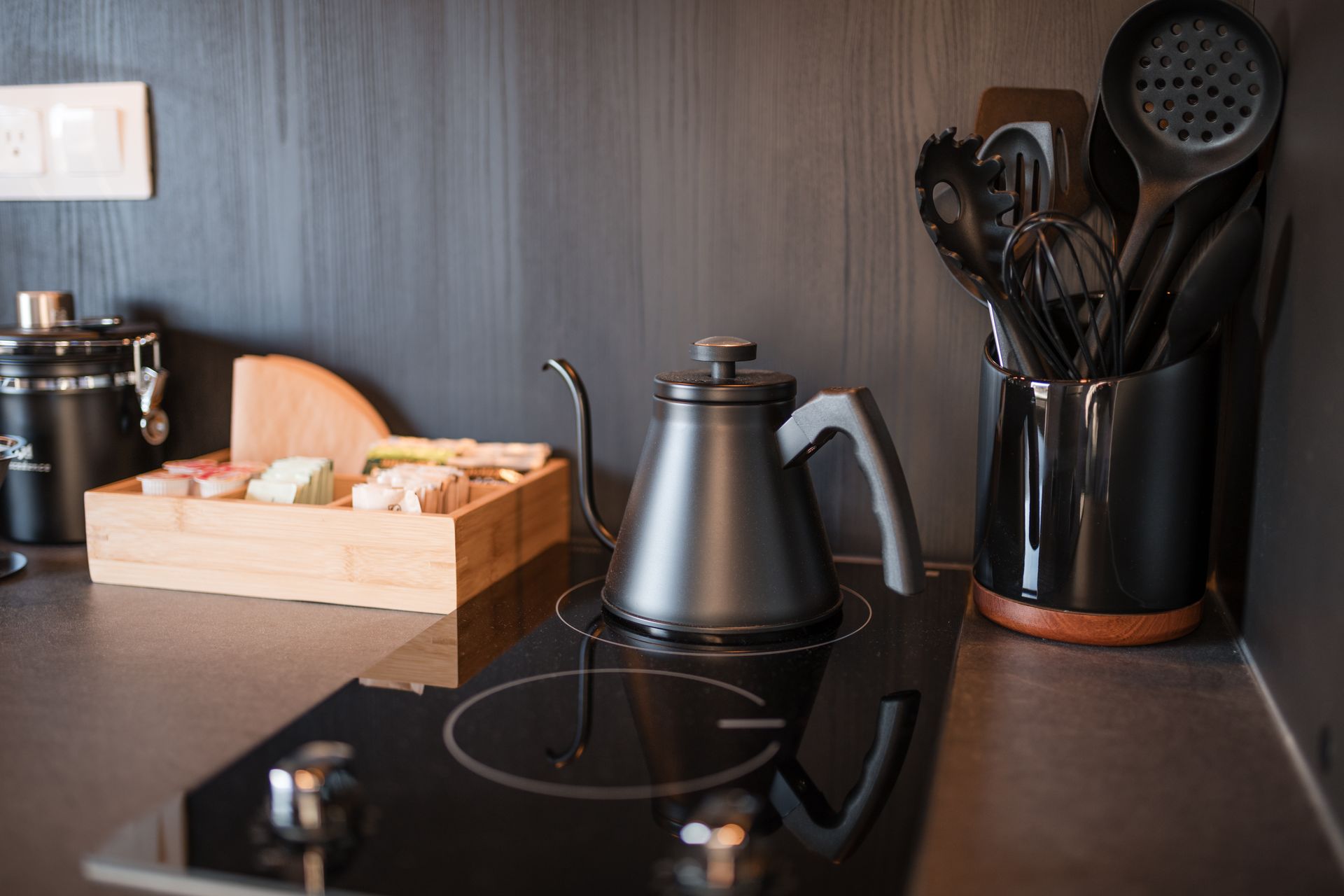 Black kettle on a stovetop, tea supplies, and utensils in a modern kitchen.