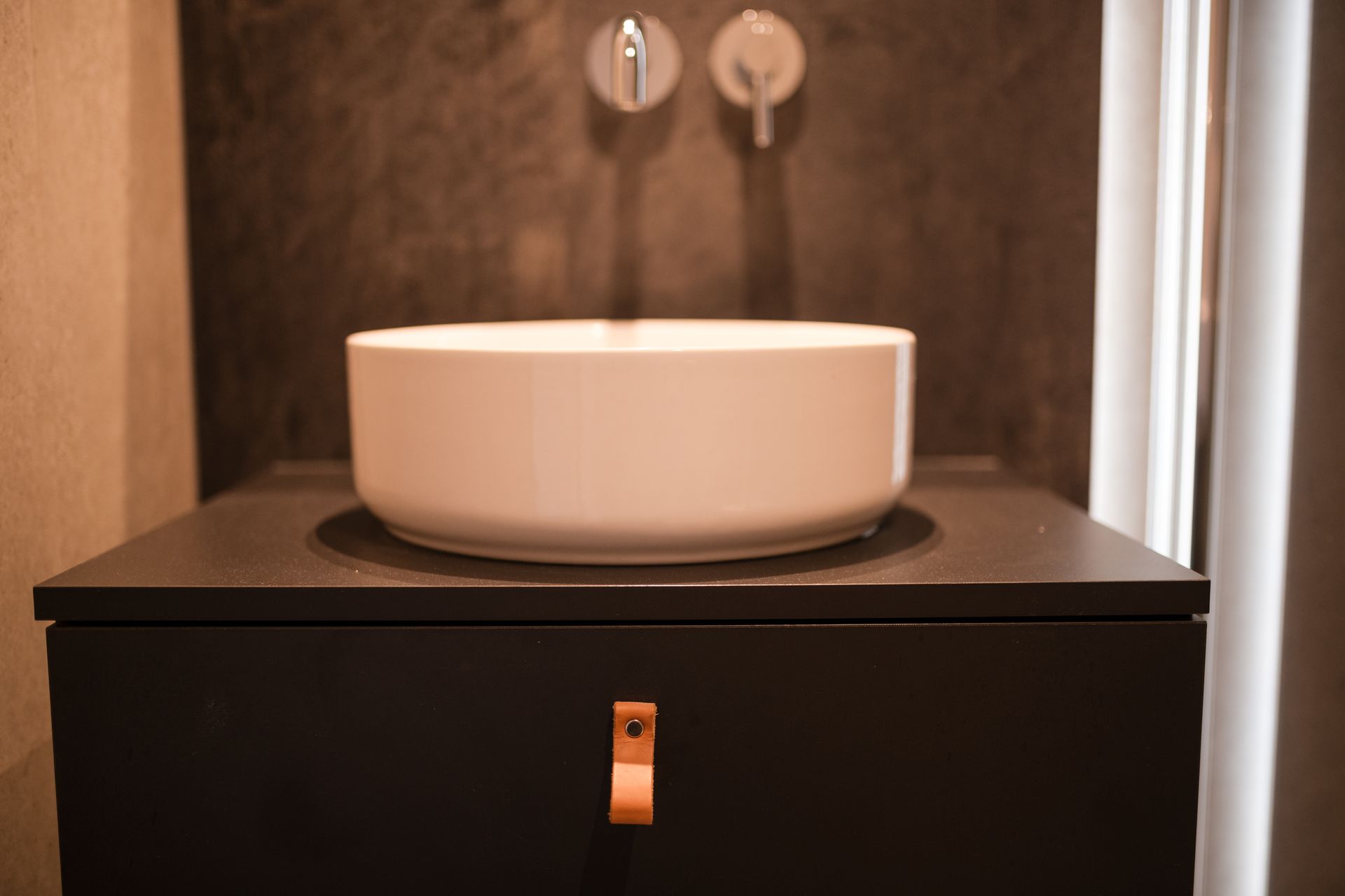 White bowl sink on dark cabinet with leather pull, against dark wall.