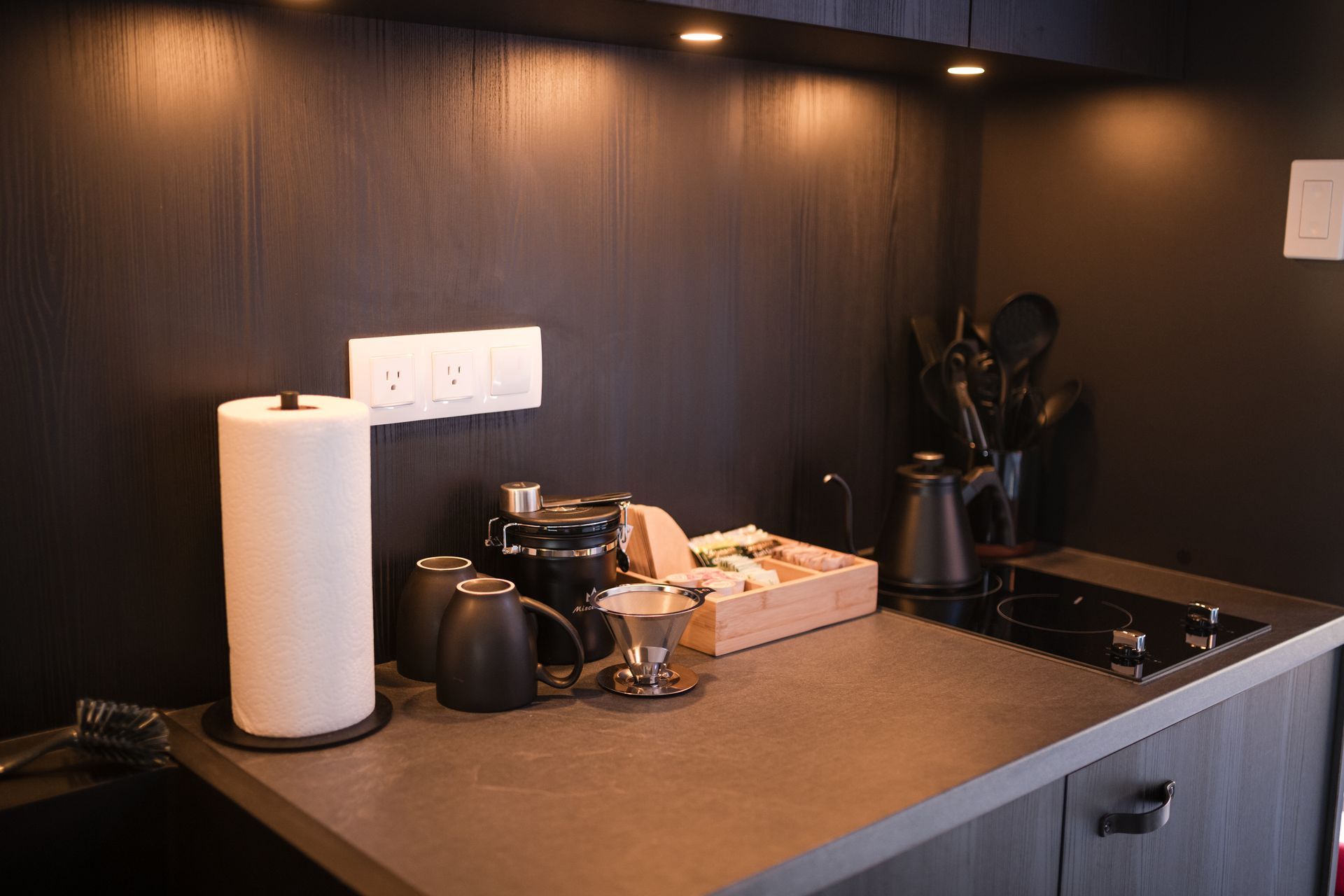 Dark kitchen countertop with coffee setup, paper towel, and overhead lighting.