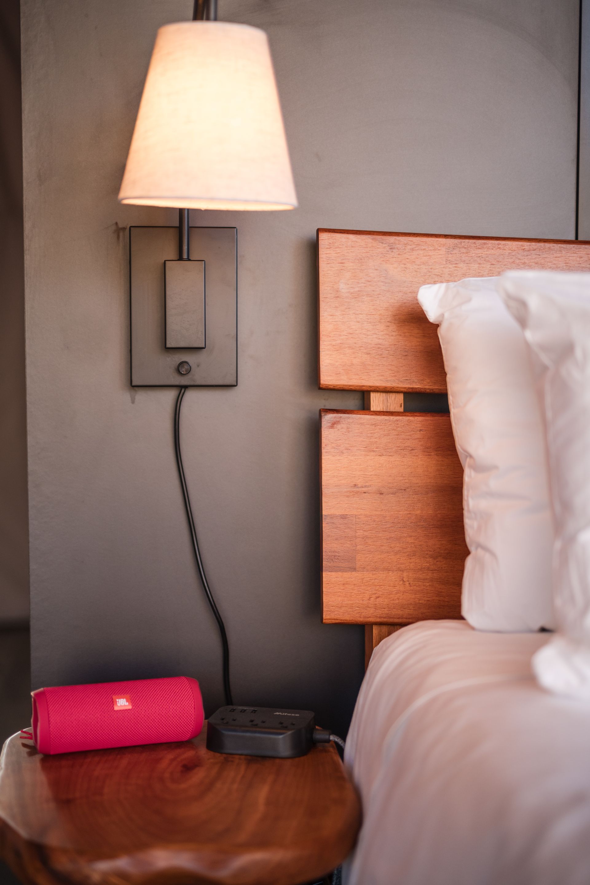 Bedside scene with lamp, wood headboard, and a pink speaker on a wooden table.