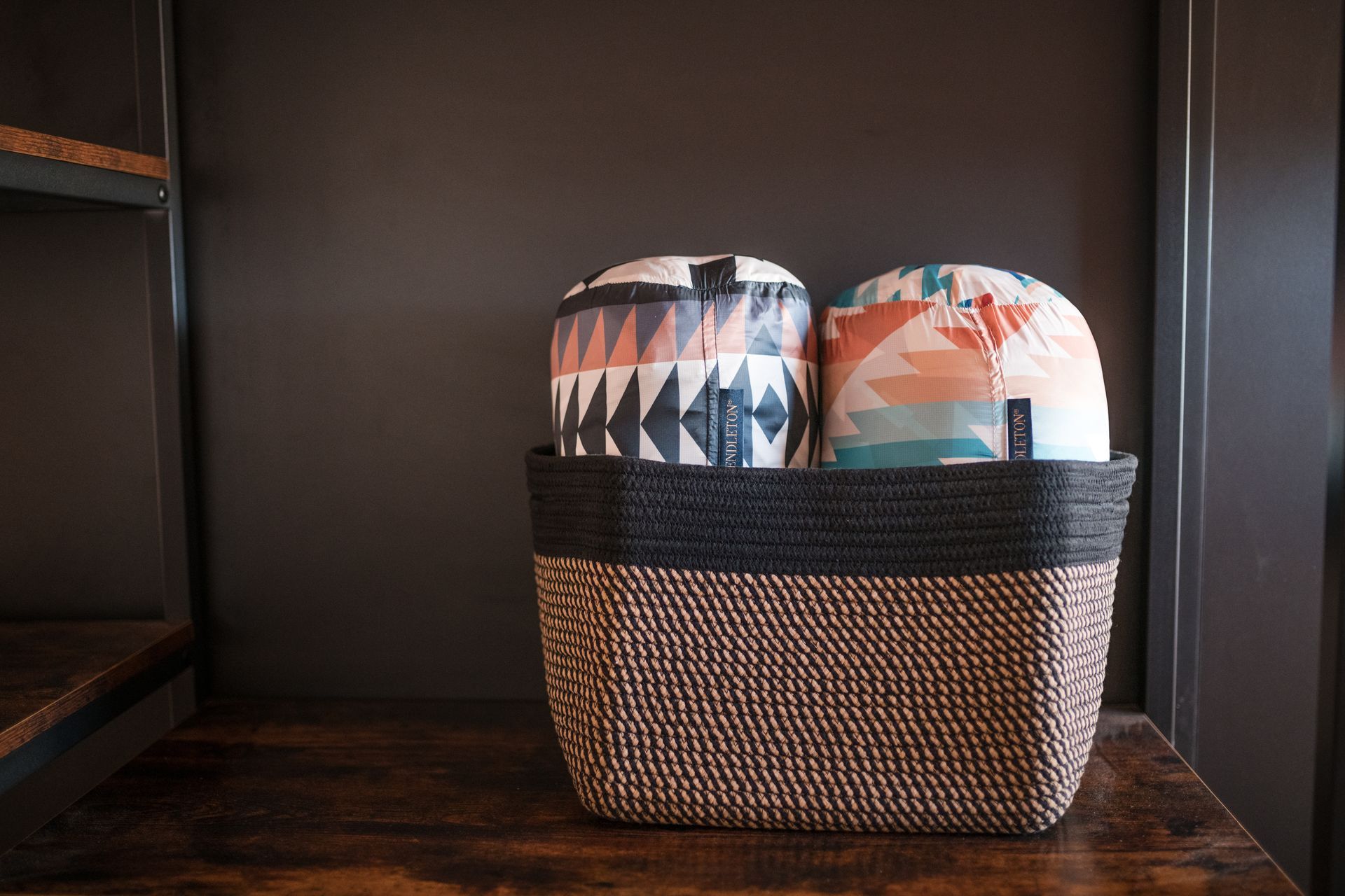 Basket holding two patterned fabric-covered items on a wooden shelf against a dark wall.