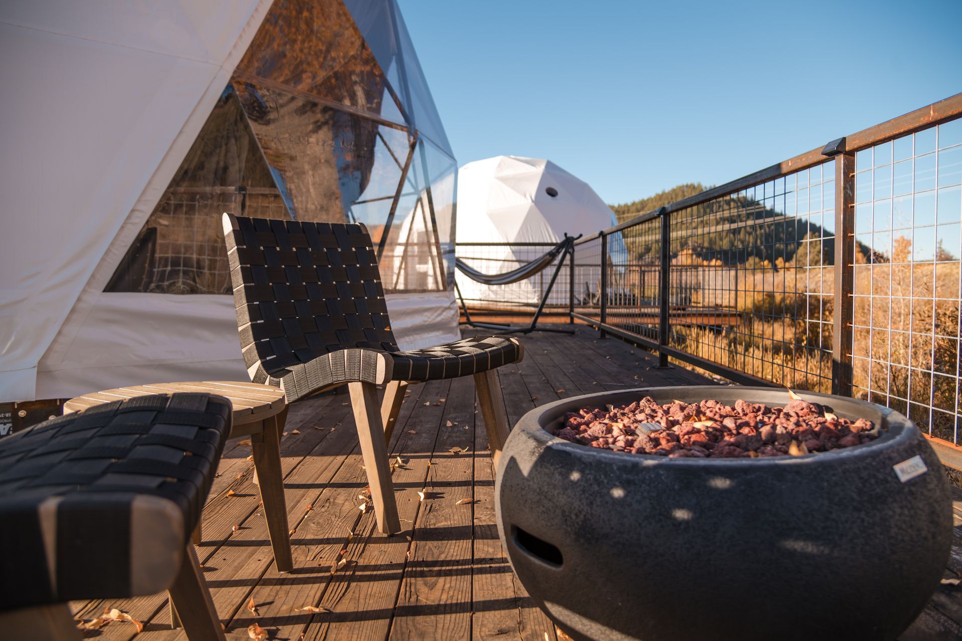 Wooden deck with chair and fire pit, with two geodesic domes in the background under a blue sky.