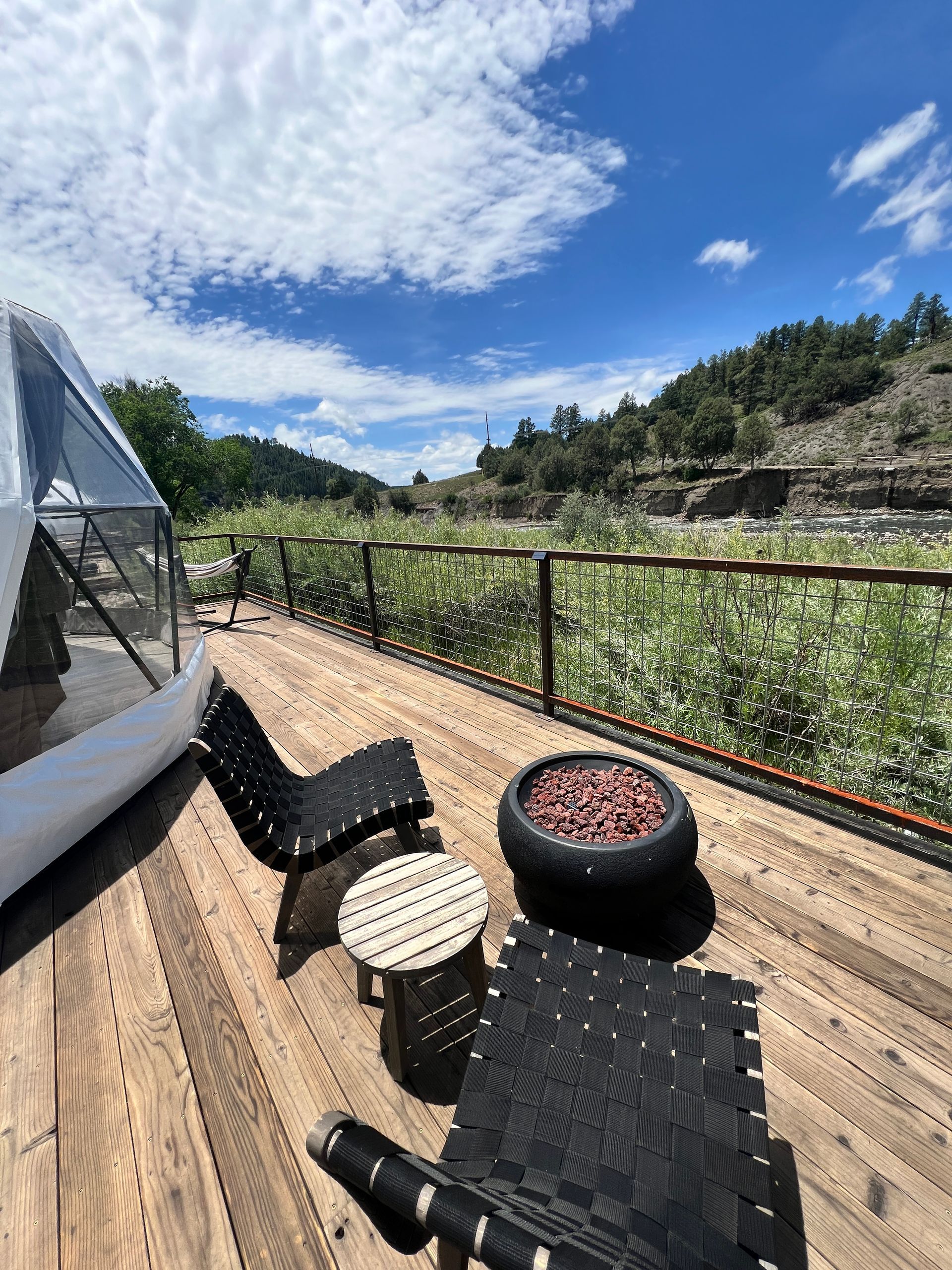 Wooden deck with chairs, a table, and potted flowers overlooks a green river with a blue sky.