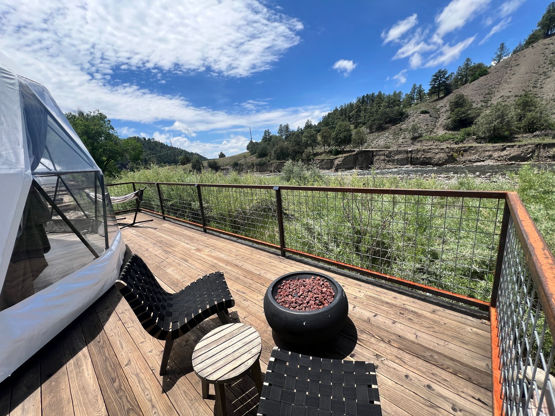 Deck with seating by a river, adjacent to a domed structure. Lush foliage and hills under a blue sky.